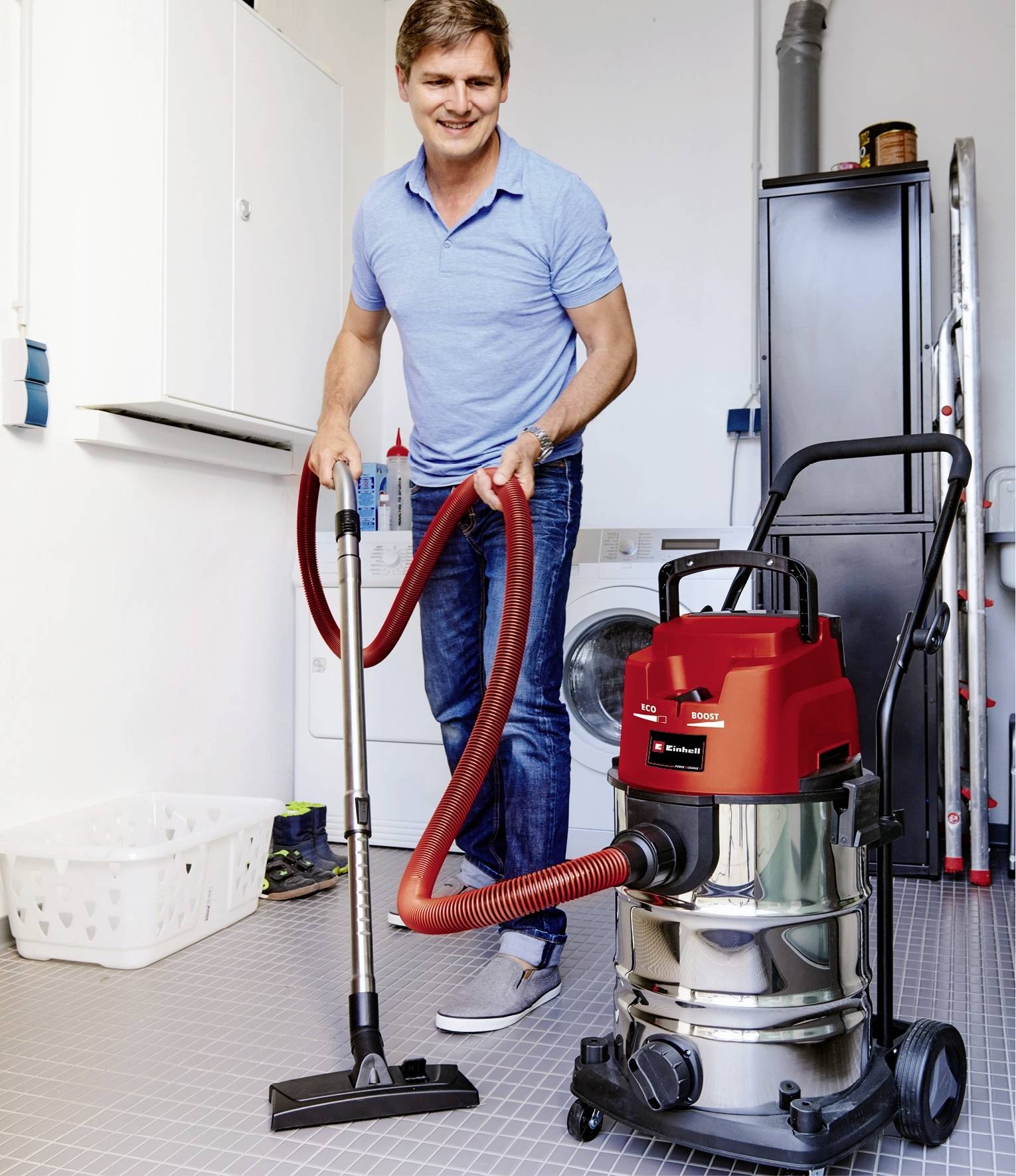 'A man in casual clothing is using a red industrial vacuum cleaner to clean a utility room with a washing machine.'