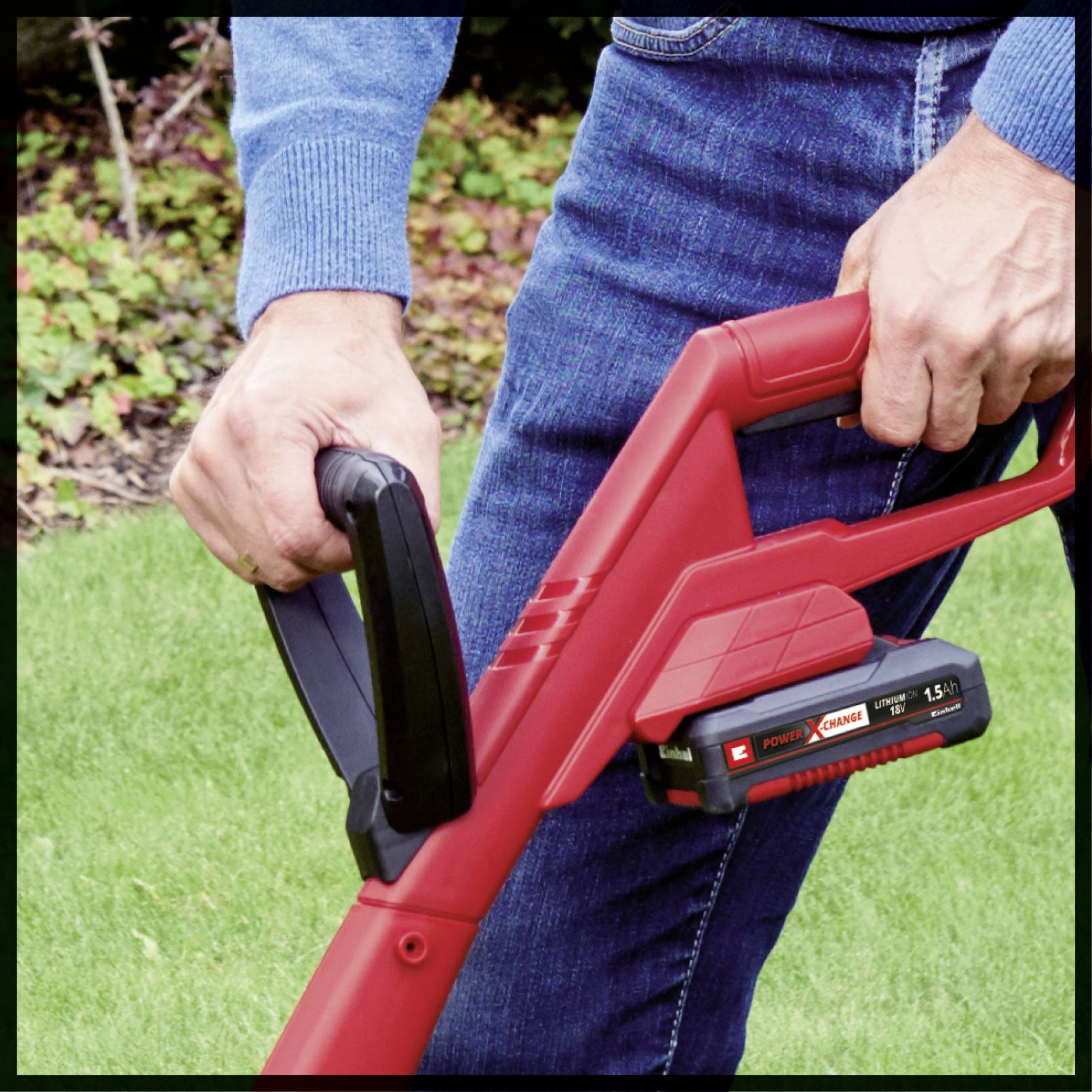 A person is holding a red lawnmower with both hands, either in use or ready for operation. Plants are visible in the background.