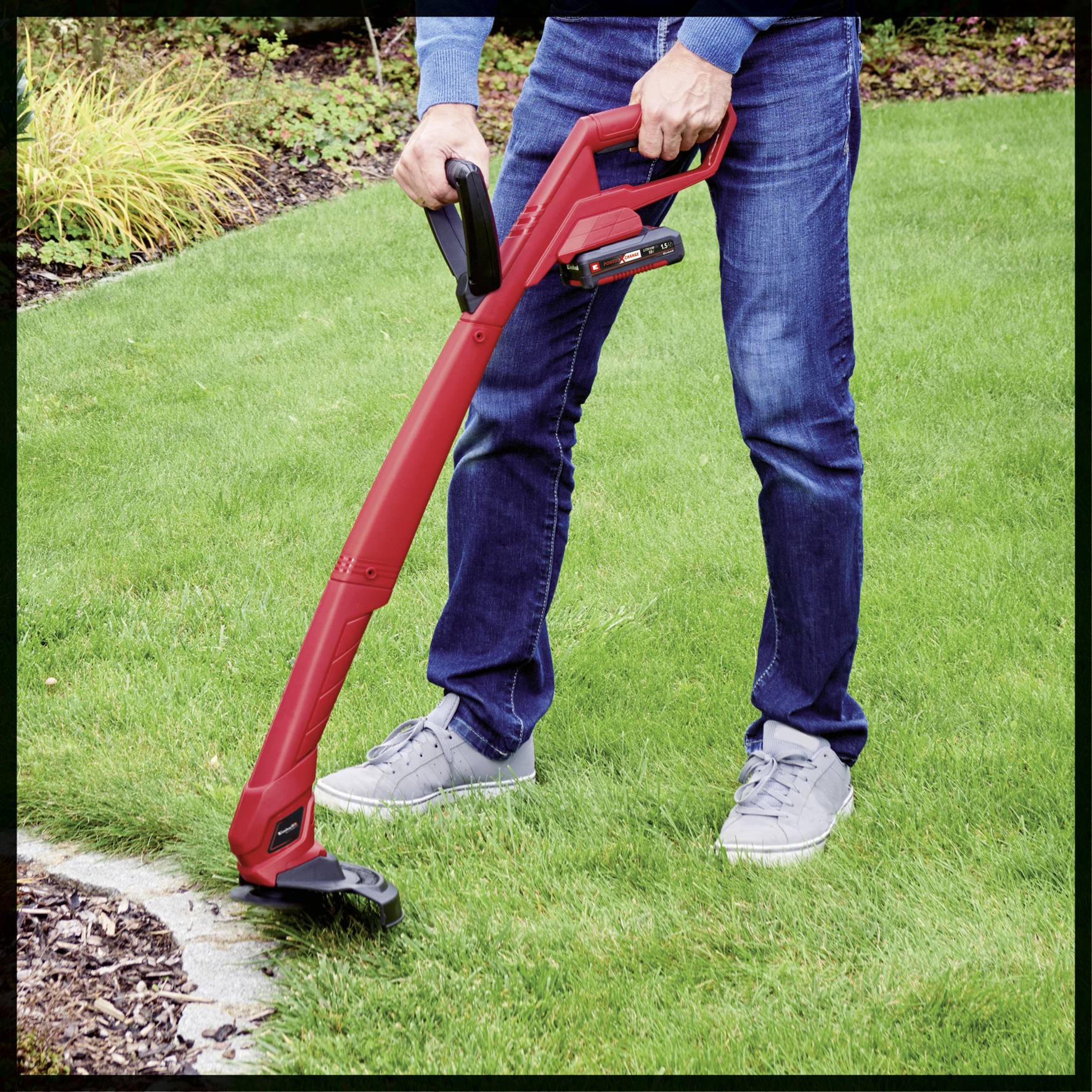 A person is cutting the lawn with a red, electrically powered lawn trimmer. The lawn is green and well-maintained.