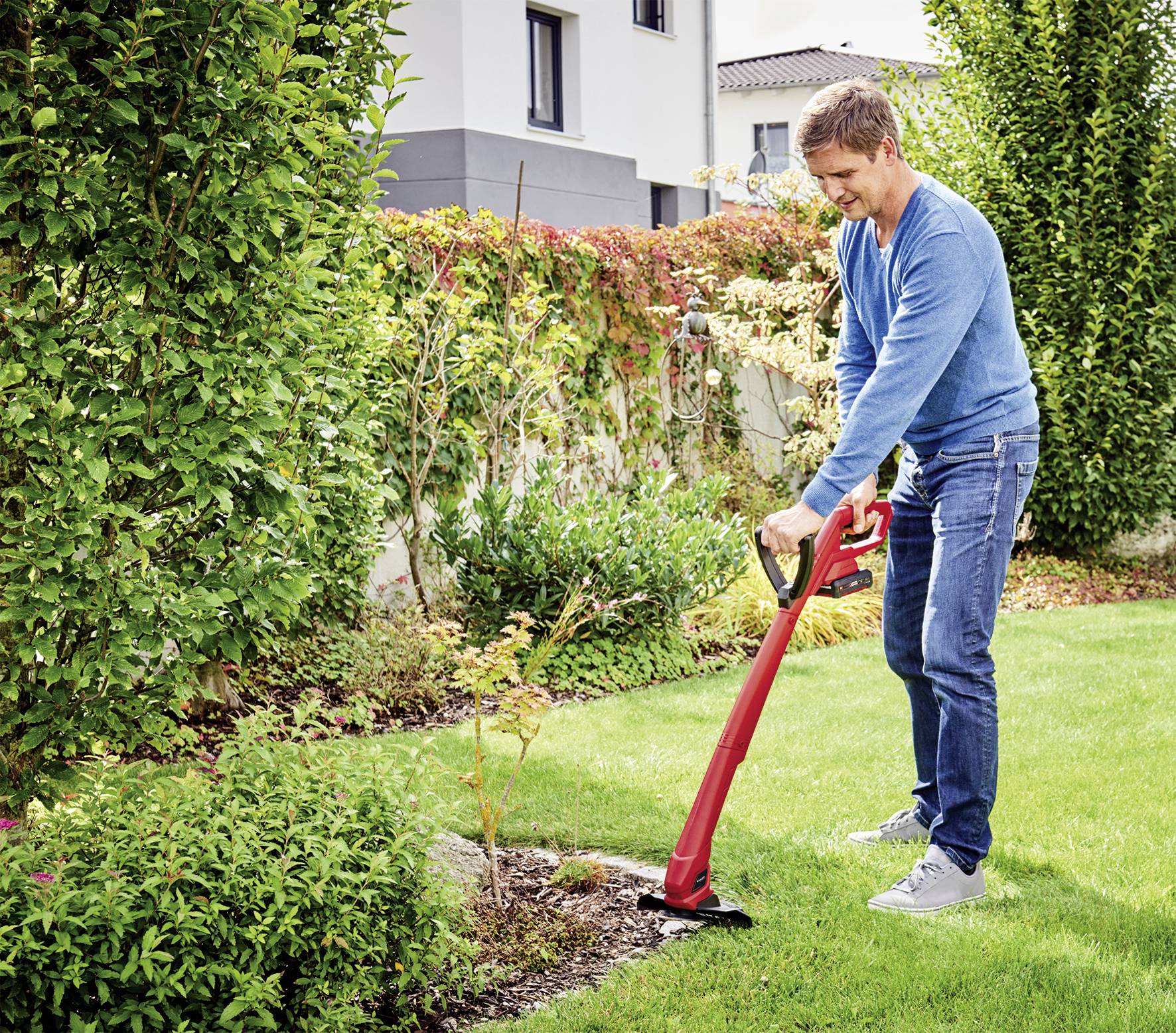 A man is mowing the lawn with a red lawn trimmer in a well-maintained garden next to a house.
