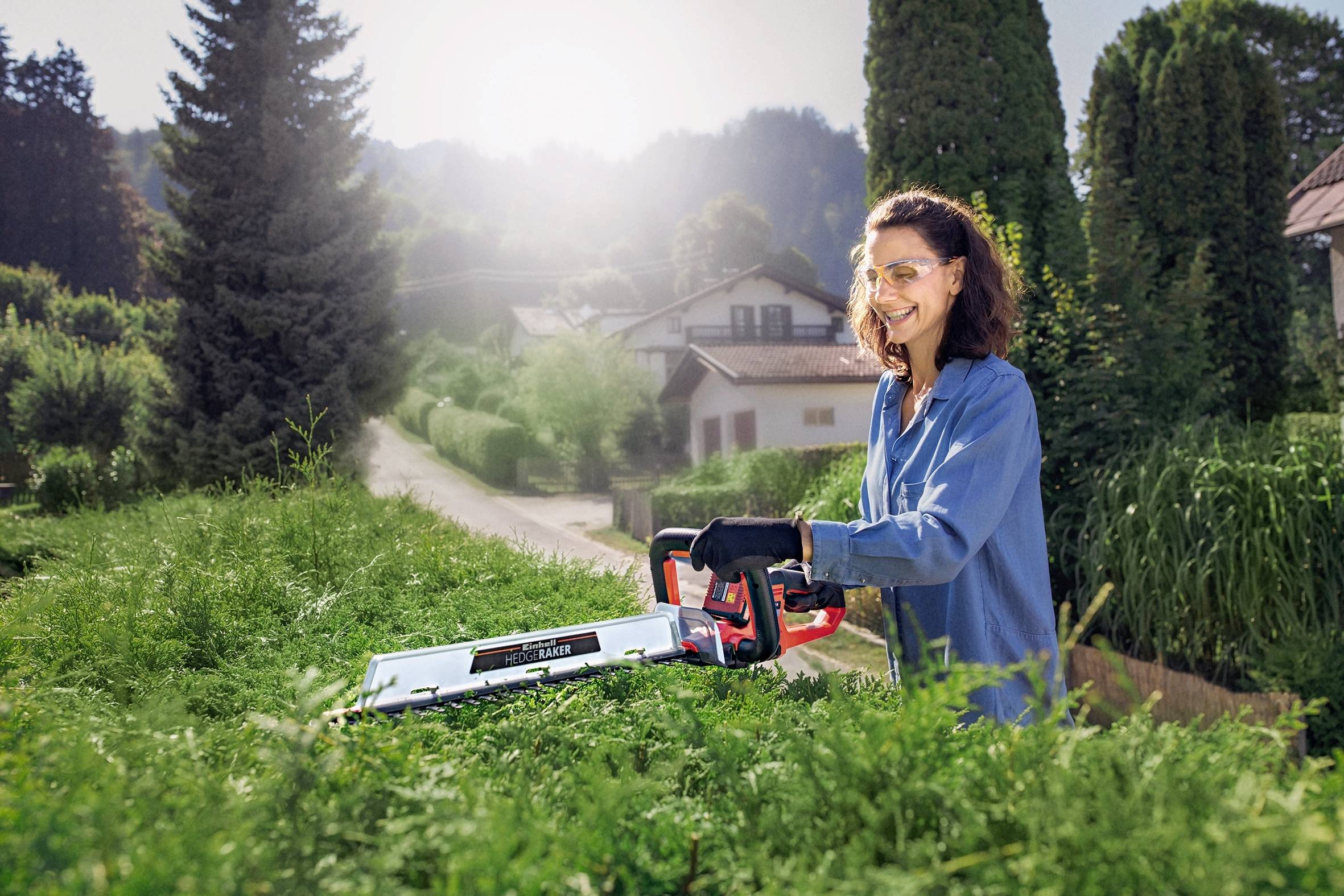 A woman is trimming a hedge in a sunny garden. She is wearing safety glasses and gloves. Trees and a house are visible in the background.