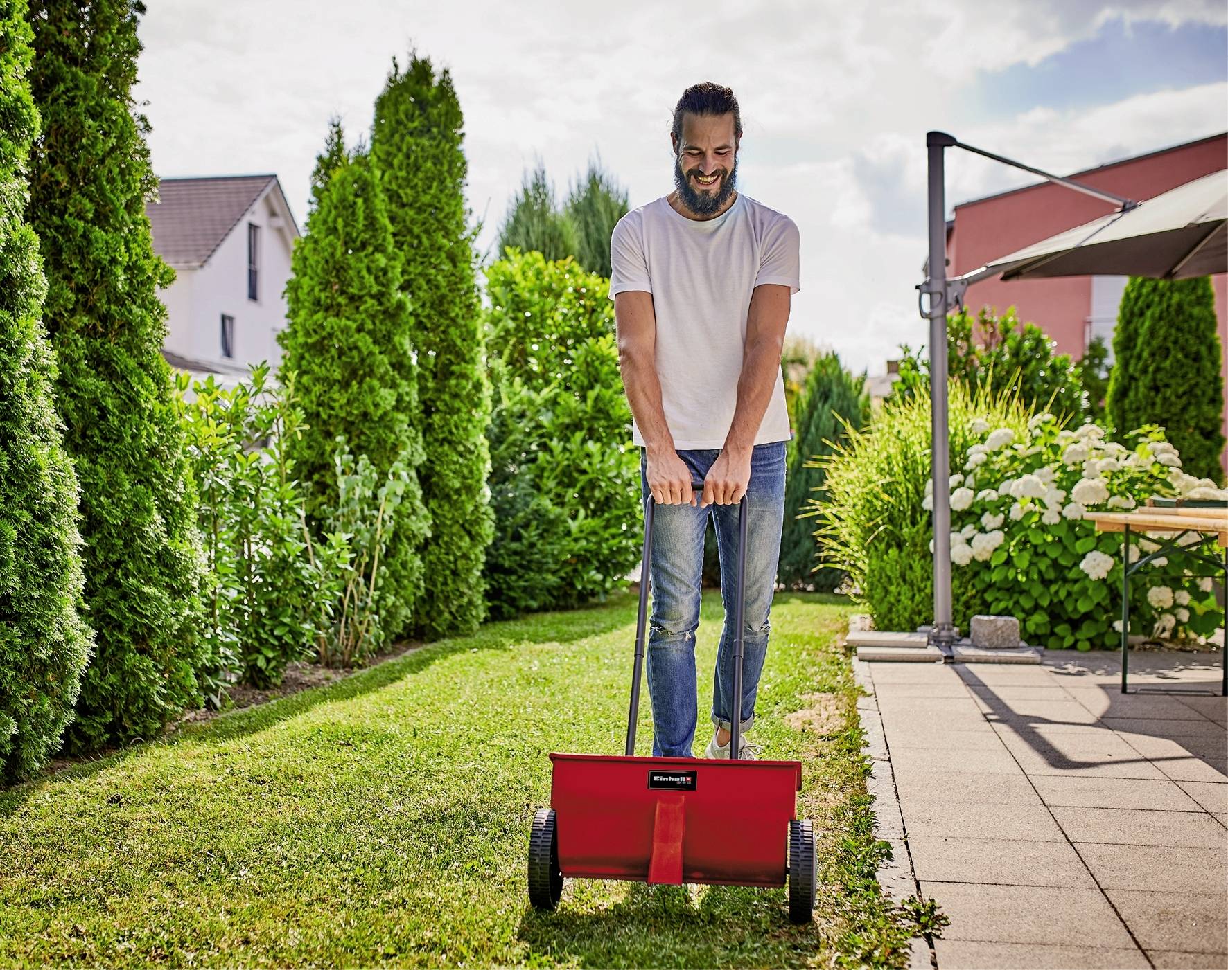 A man is mowing the lawn with a hand mower on a sunny day in a garden. Trees and a house are visible in the background.
