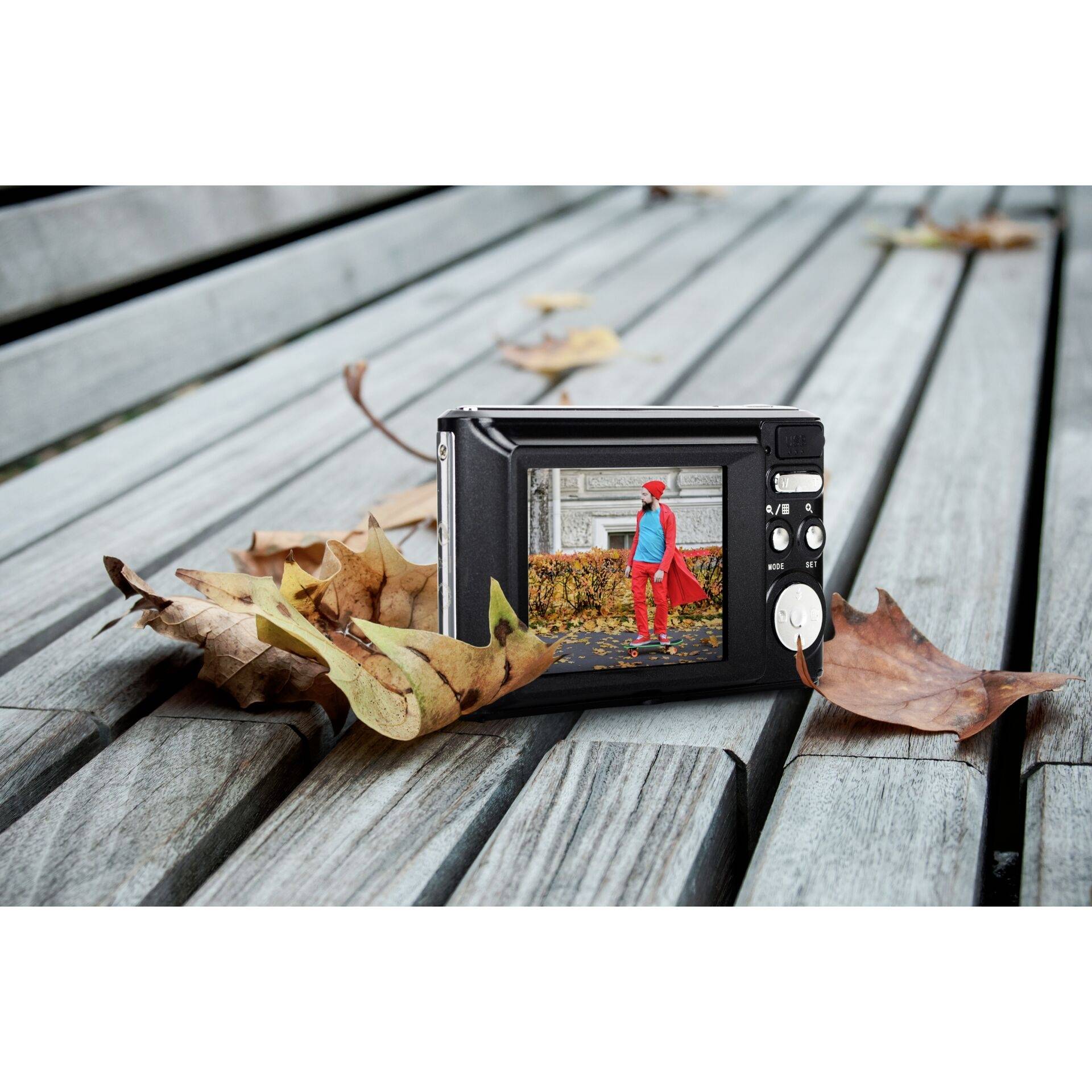 A digital camera is lying on a wooden table. On the screen, a person wearing a red jacket and hat can be seen walking along a park path.