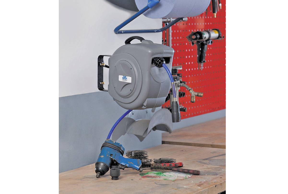 An automatic hose reel mounted on the wall, connected to pneumatic tools and brushes on a workbench in the foreground.