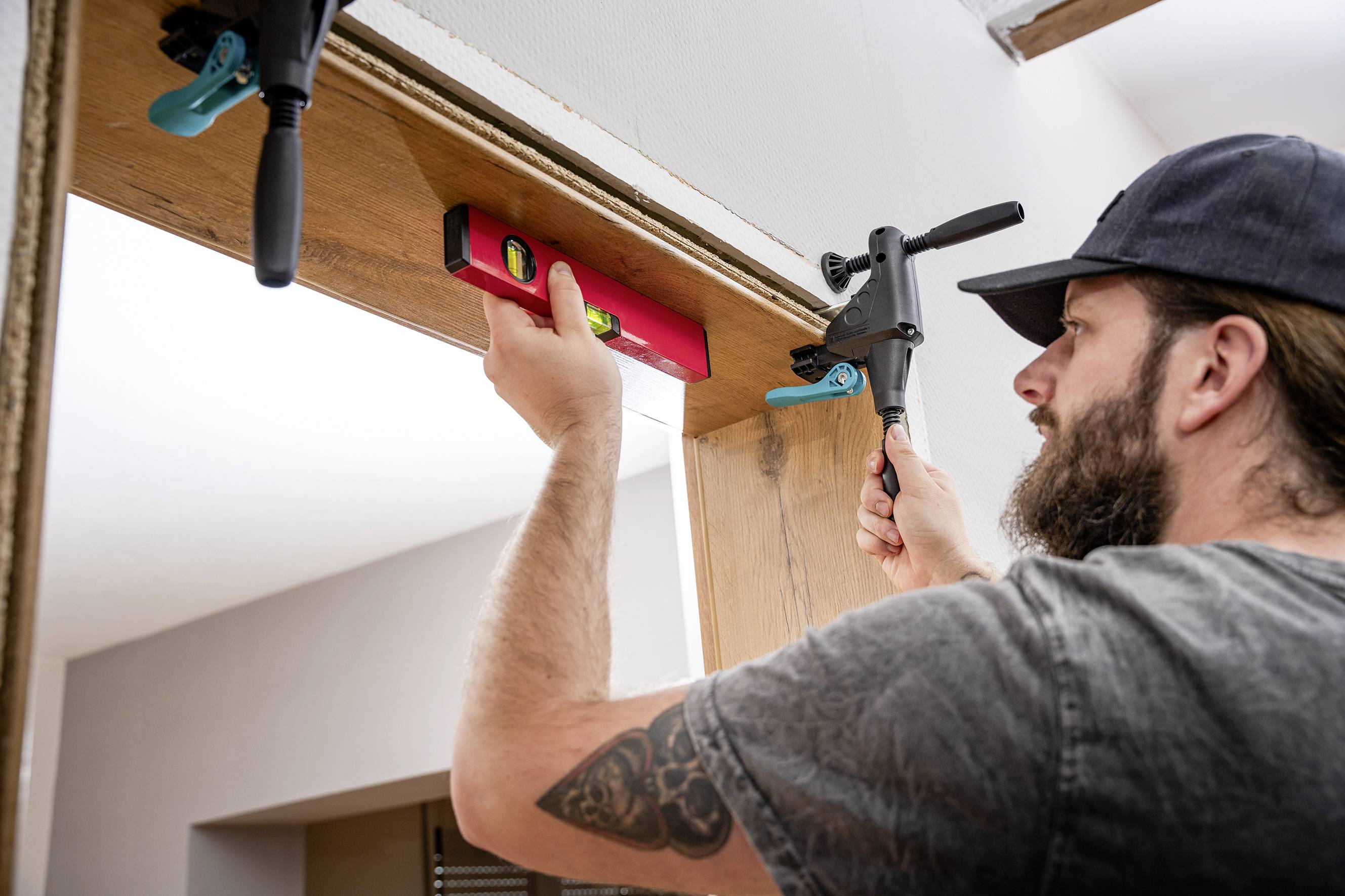 'A man aligns a wooden door with a red spirit level and uses a clamp to secure the wood. He is wearing a grey T-shirt and a black cap.'