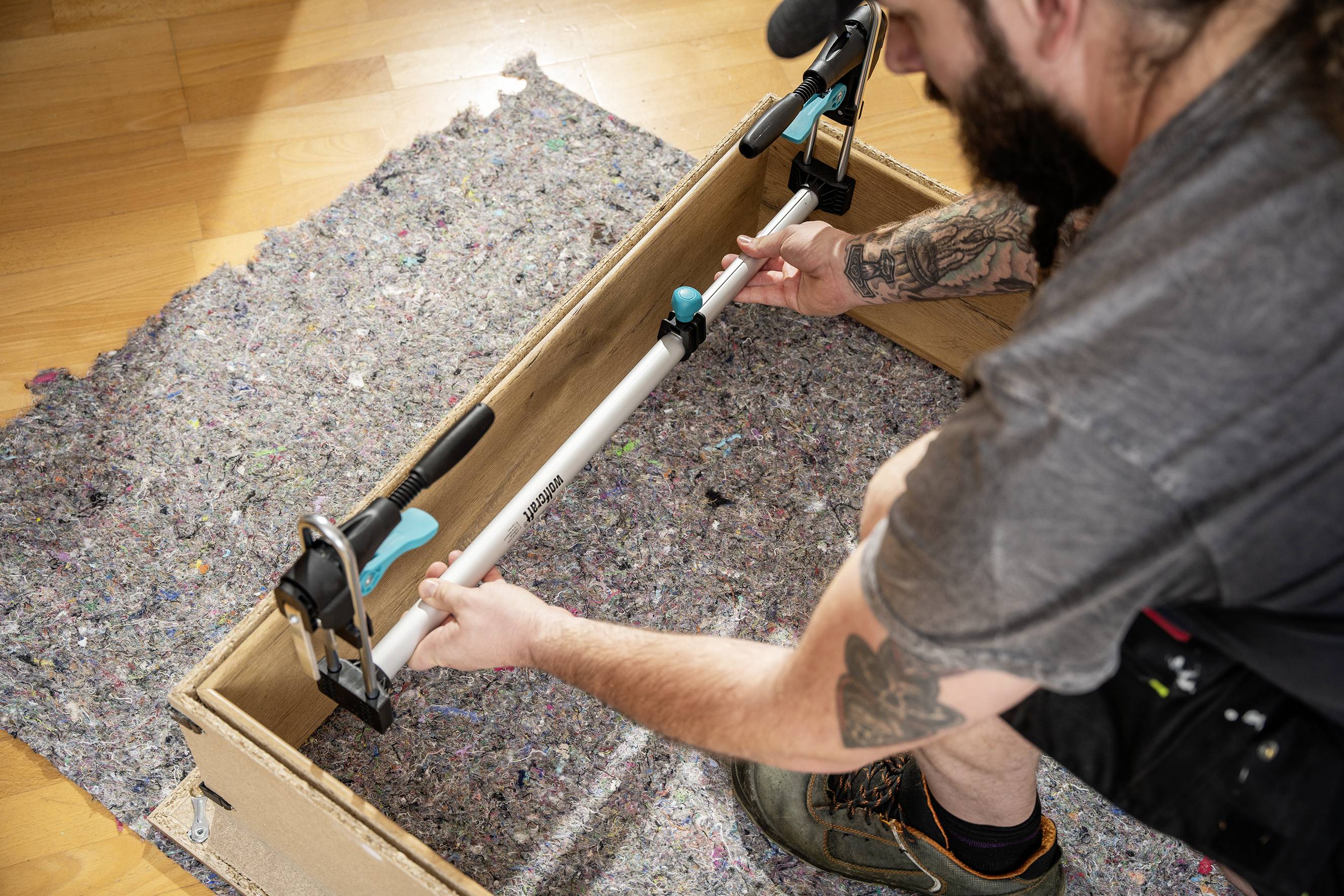 A person is mounting a pipe with tools on a board. A carpet is laid on the floor. They are wearing a grey T-shirt and have tattooed arms.