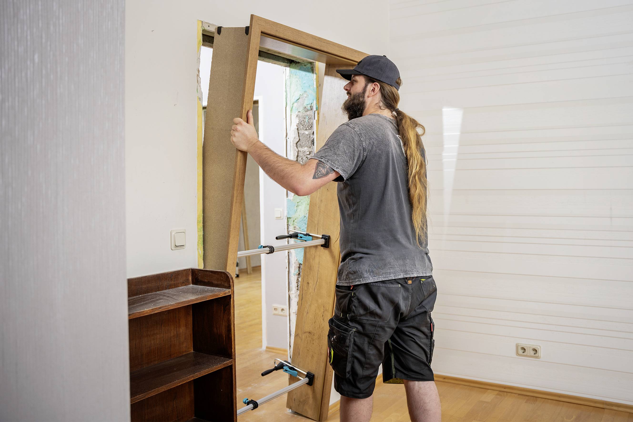 A man with long hair is installing a door inside a room. He is wearing work clothes and a cap.
