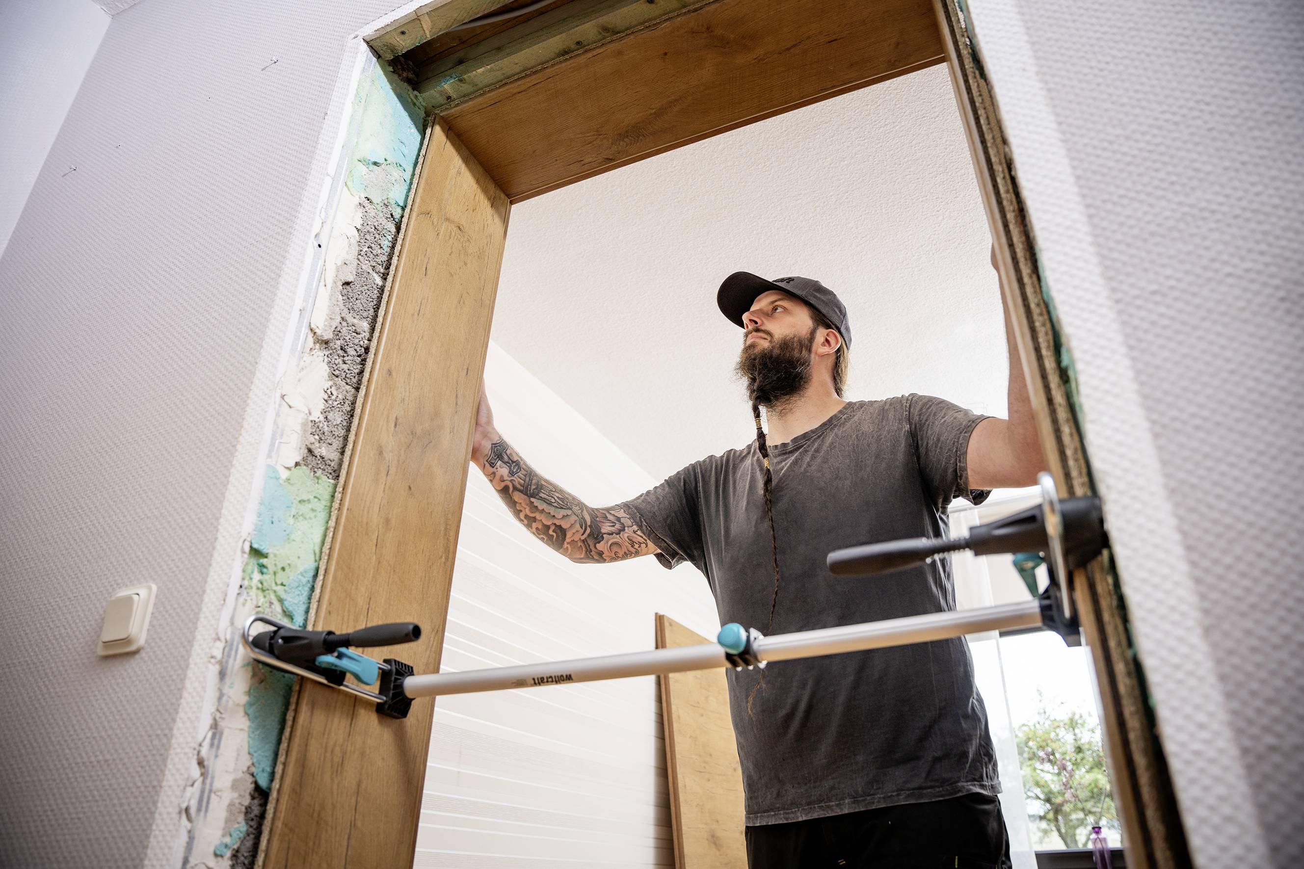 A man with tattoos is installing a wooden door in an interior space, holding it steady with a clamping tool.