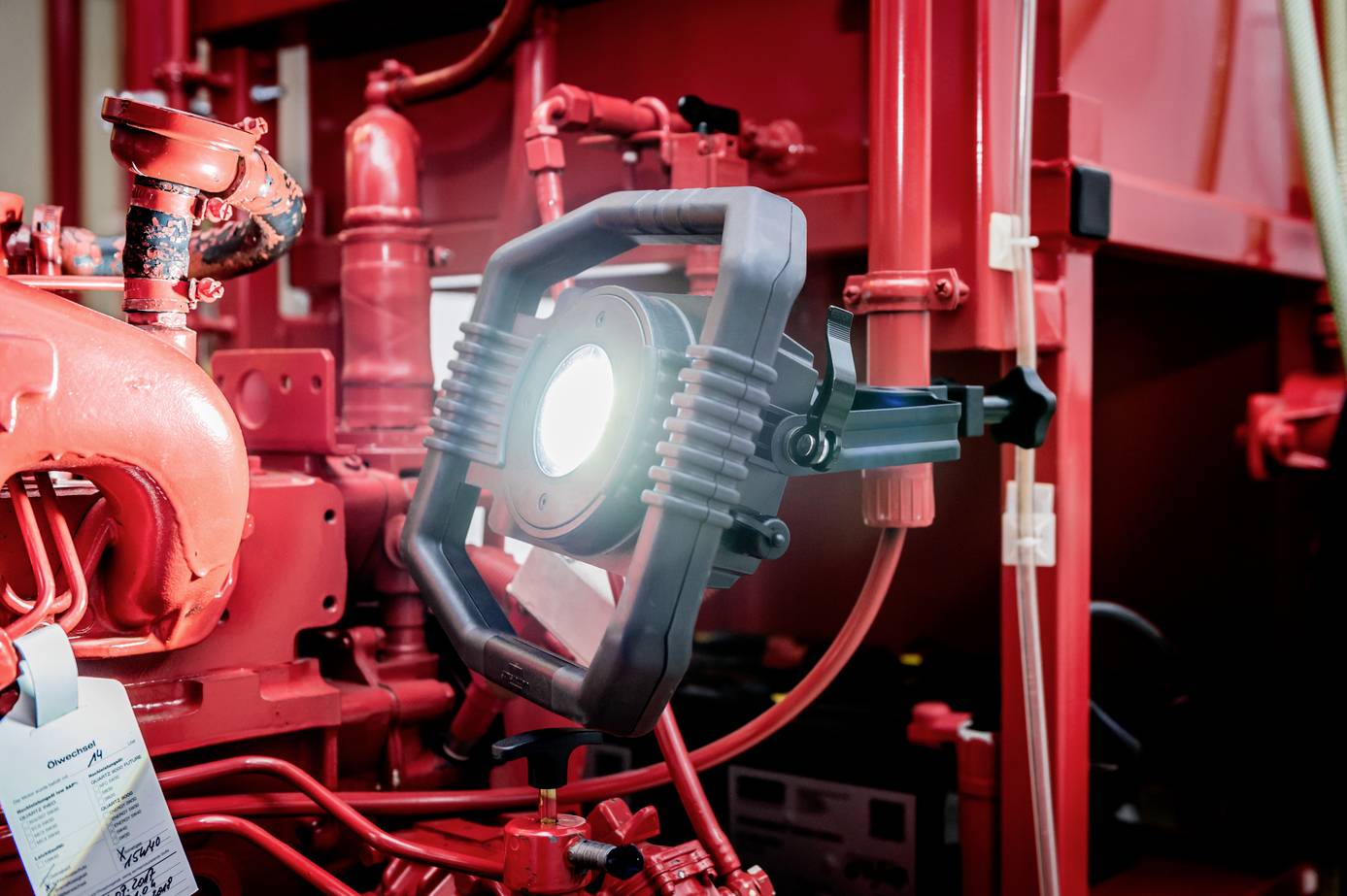 A workshop light illuminates the engine compartment of a machine. The scene is surrounded by red machine parts and pipes.