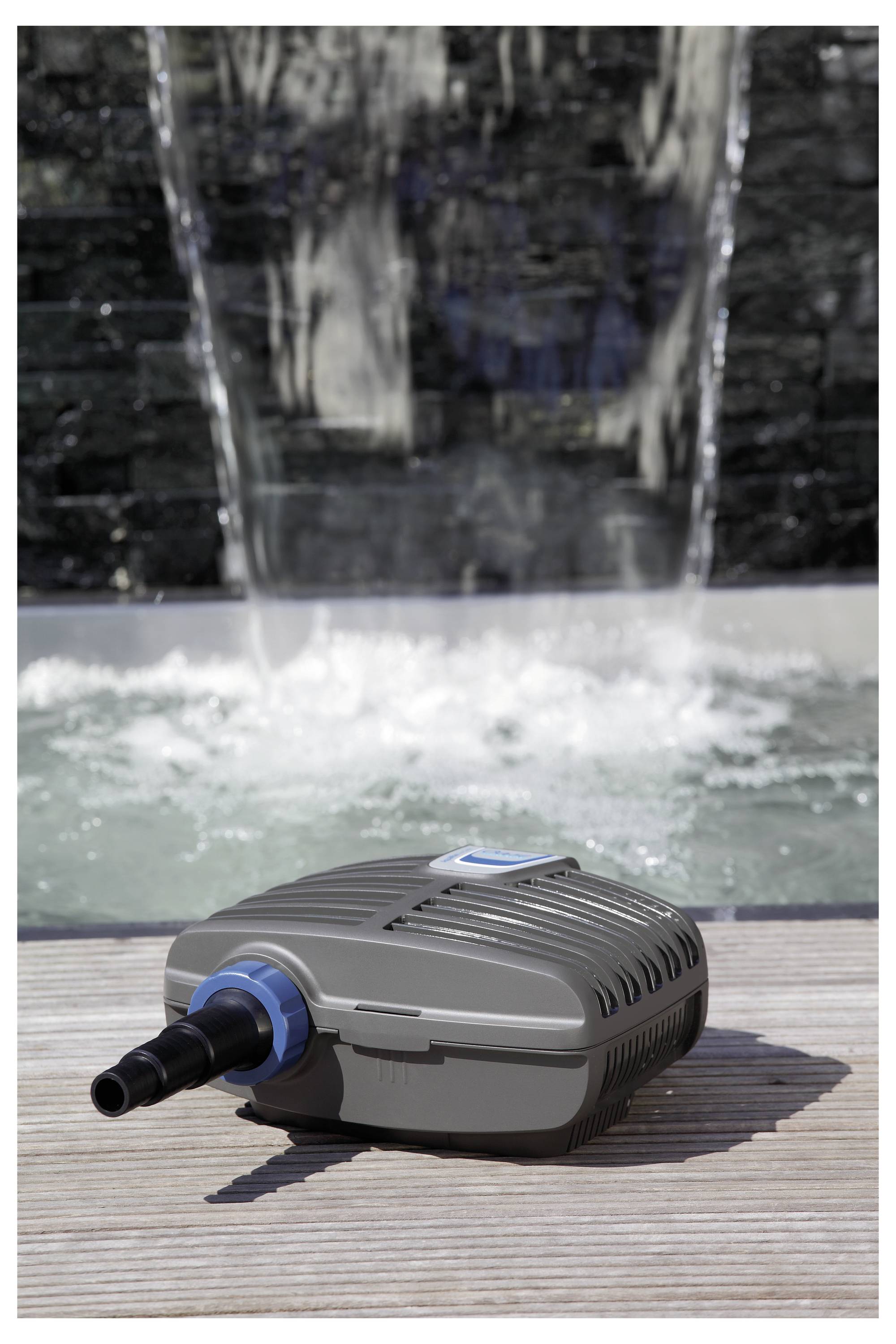 A gray water pump lies on a wooden deck in front of a flowing waterfall.