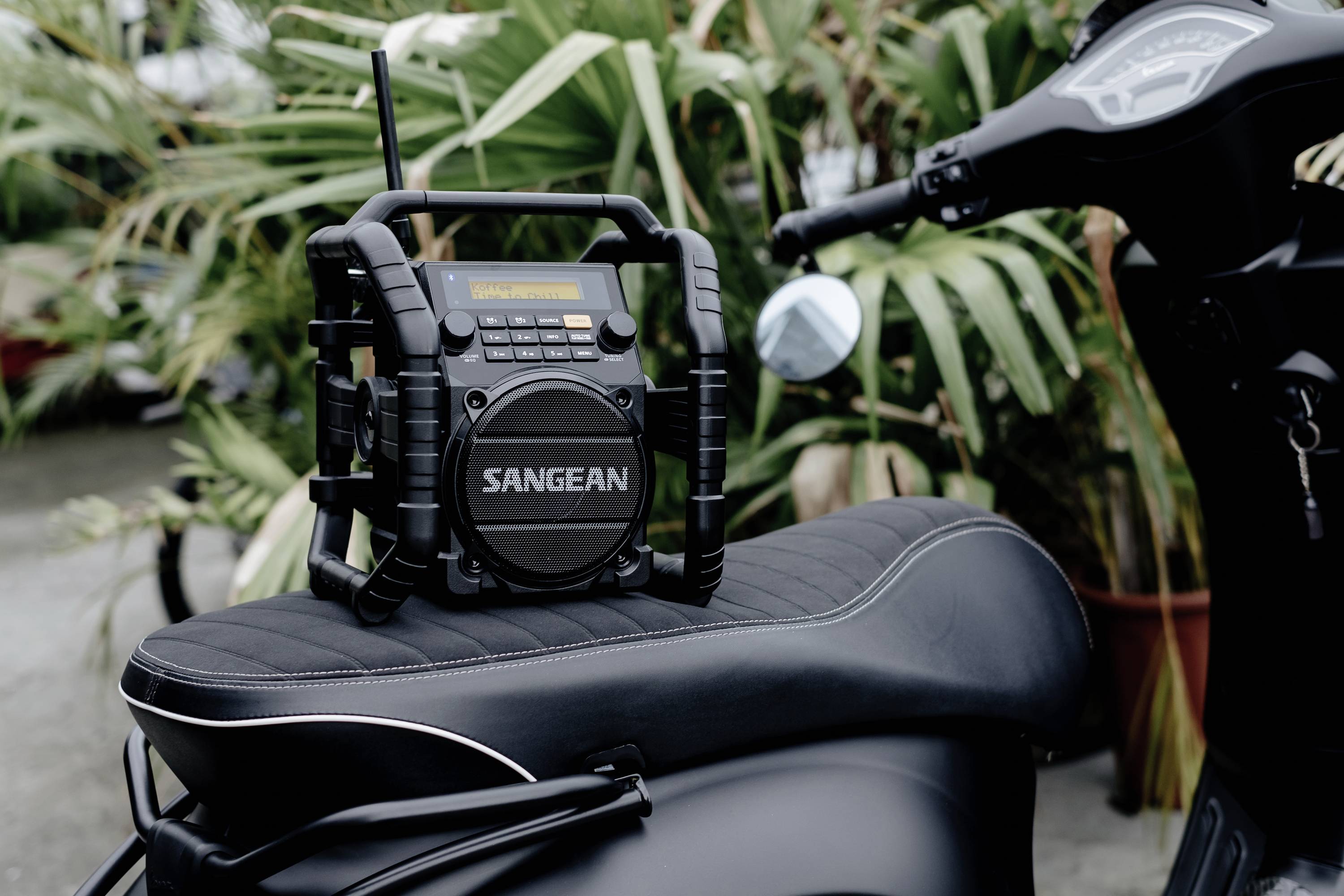 A portable radio with the label 'Sangean' sits on a motorcycle seat. Plants can be seen in the background.