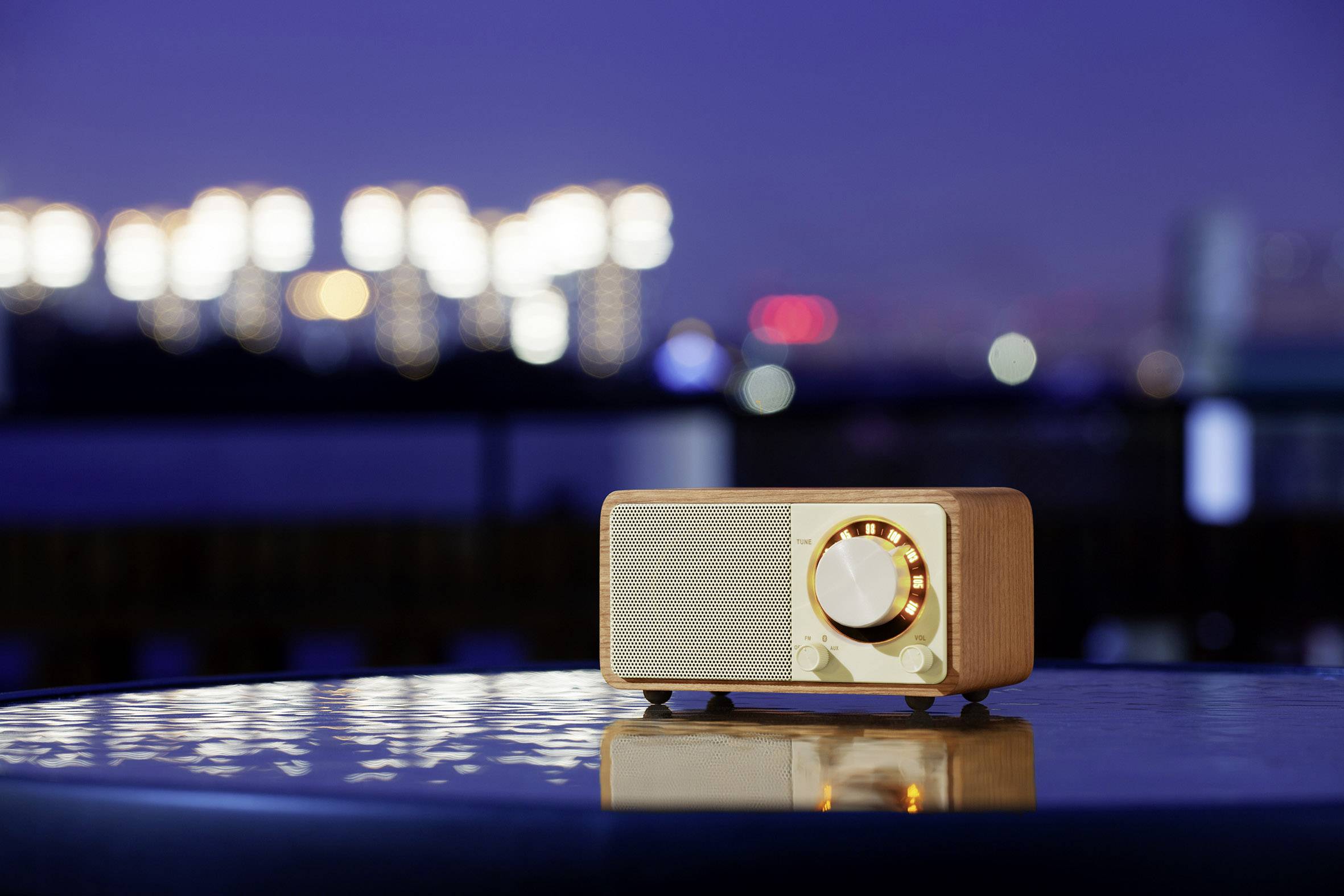 A vintage radio sits at night on a smooth table outdoors, illuminated by the blurred lights of a city in the background.