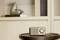 A small retro radio on a wooden table in front of a white shelf with a decorative vase in the background.