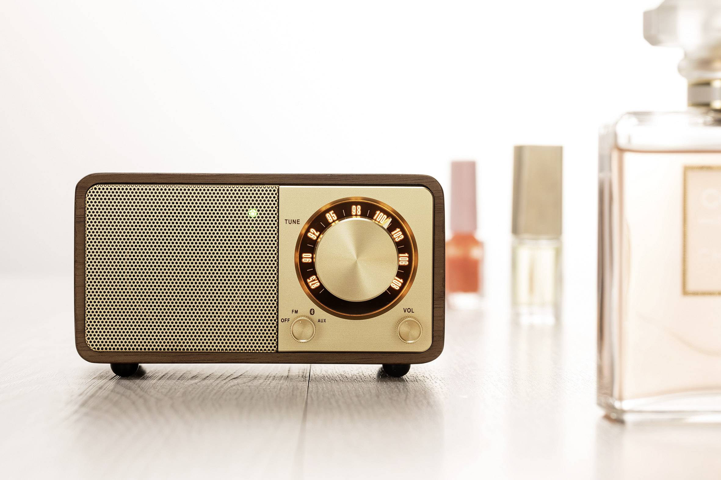 A vintage radio with an illuminated rotary dial sits on a table. Portions of perfume bottles can be seen in the background.