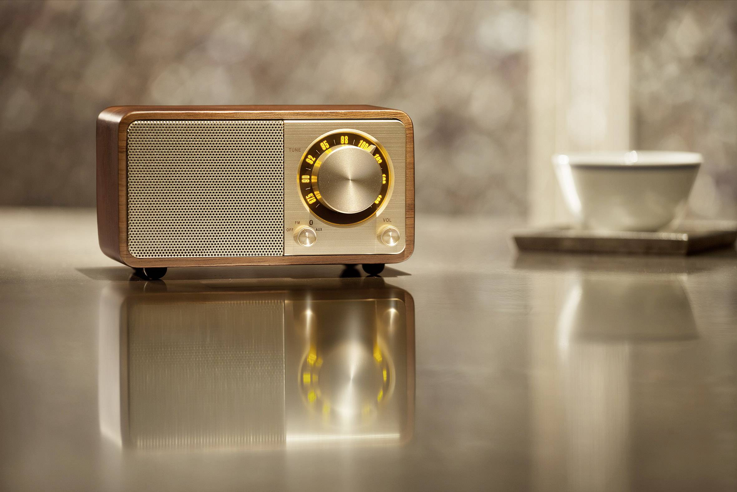 A small retro radio with a wooden casing and golden knobs sits on a glossy table. Beside it is a white cup.