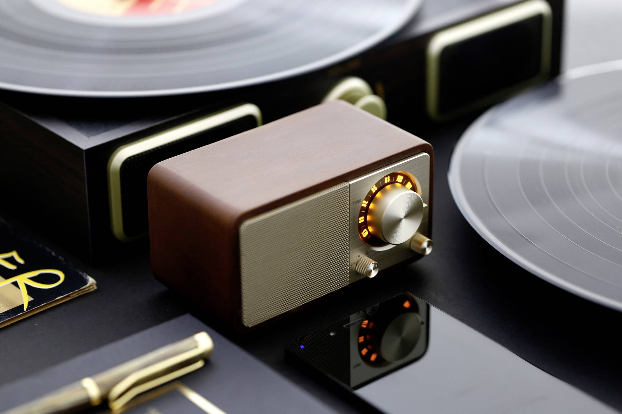 Small, elegant speaker in a retro design with a wooden enclosure and illuminated volume control, surrounded by vinyl records.
