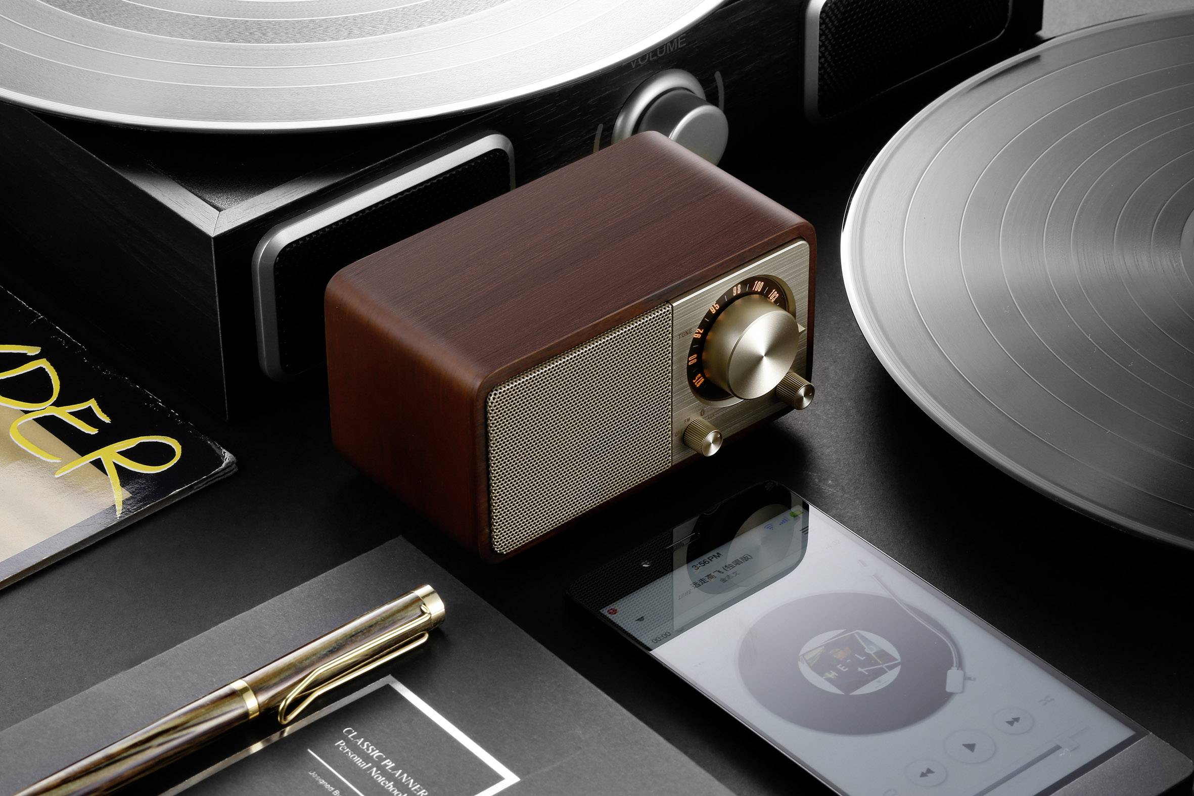 A small, vintage-inspired radio sits on a table beside vinyl records, a pen, and a smartphone displaying music.