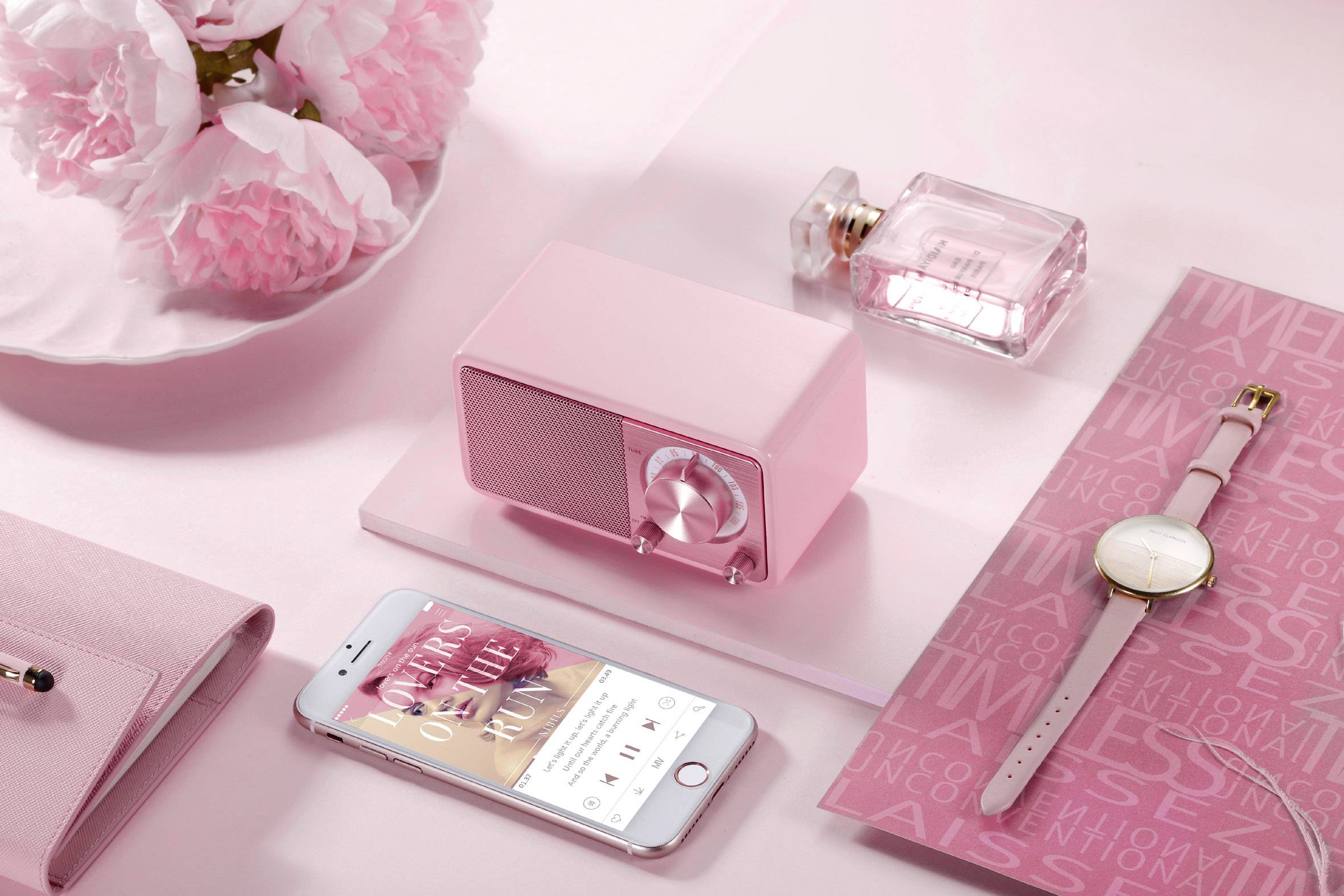 On a pink background, there is a small pink radio, a perfume bottle, a smartphone with an open music app, a wristwatch, and a notebook.