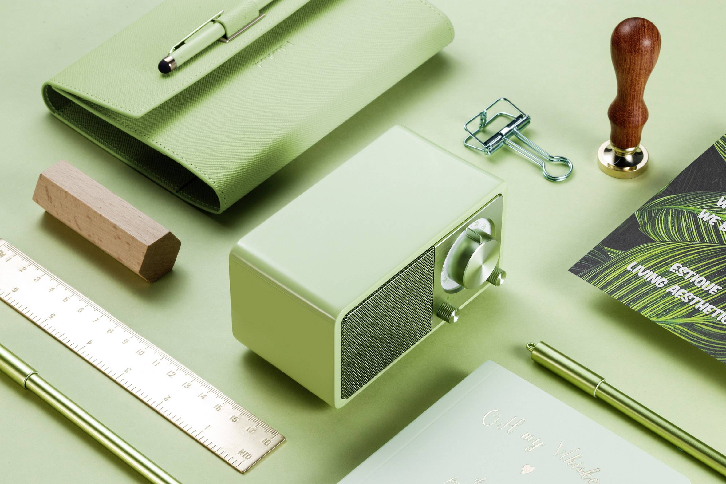 Green desk accessories: Radio, stamp, paperclip, notebook and pens on a green background, arranged in a stylish layout.