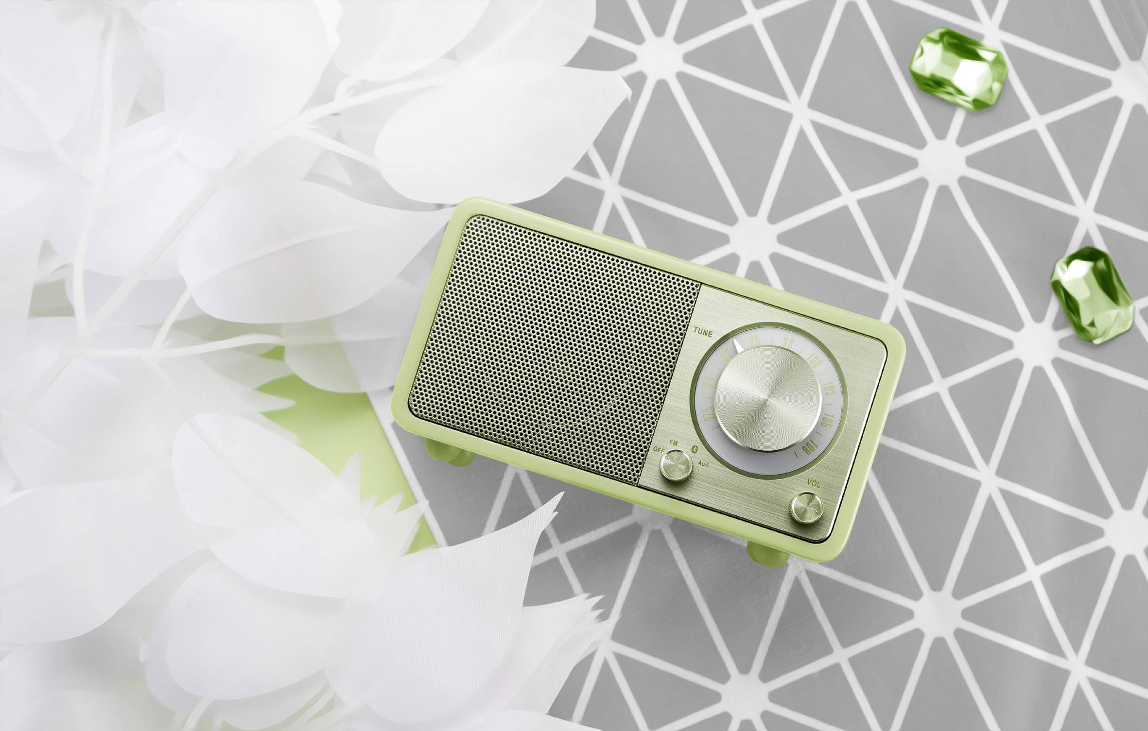 A small, green radio with a speaker and rotary knobs sits on a grey, geometric pattern. Next to it lie green gemstones.