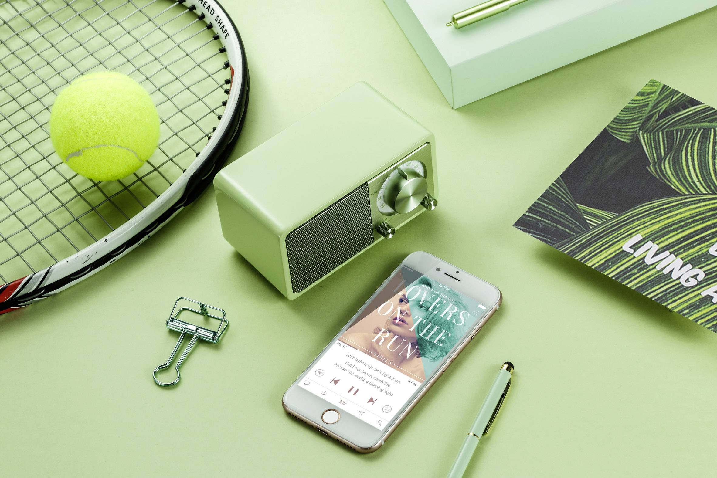 Tennis ball, racket, radio, mobile phone with music app, and notebook arranged on a green table.