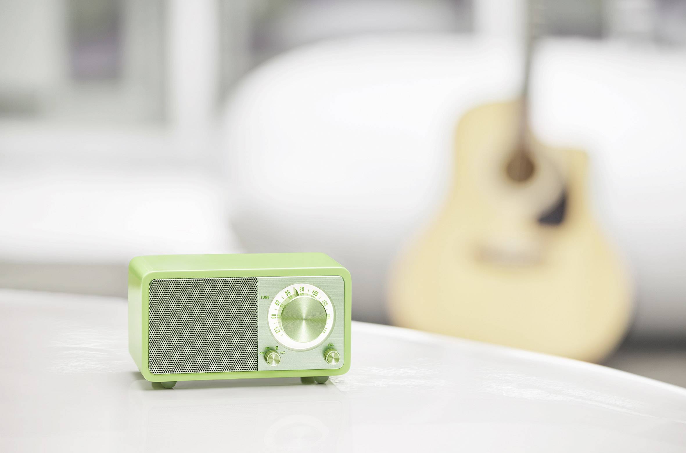A green radio sits on a white table. In the background, an acoustic guitar is blurrily visible.