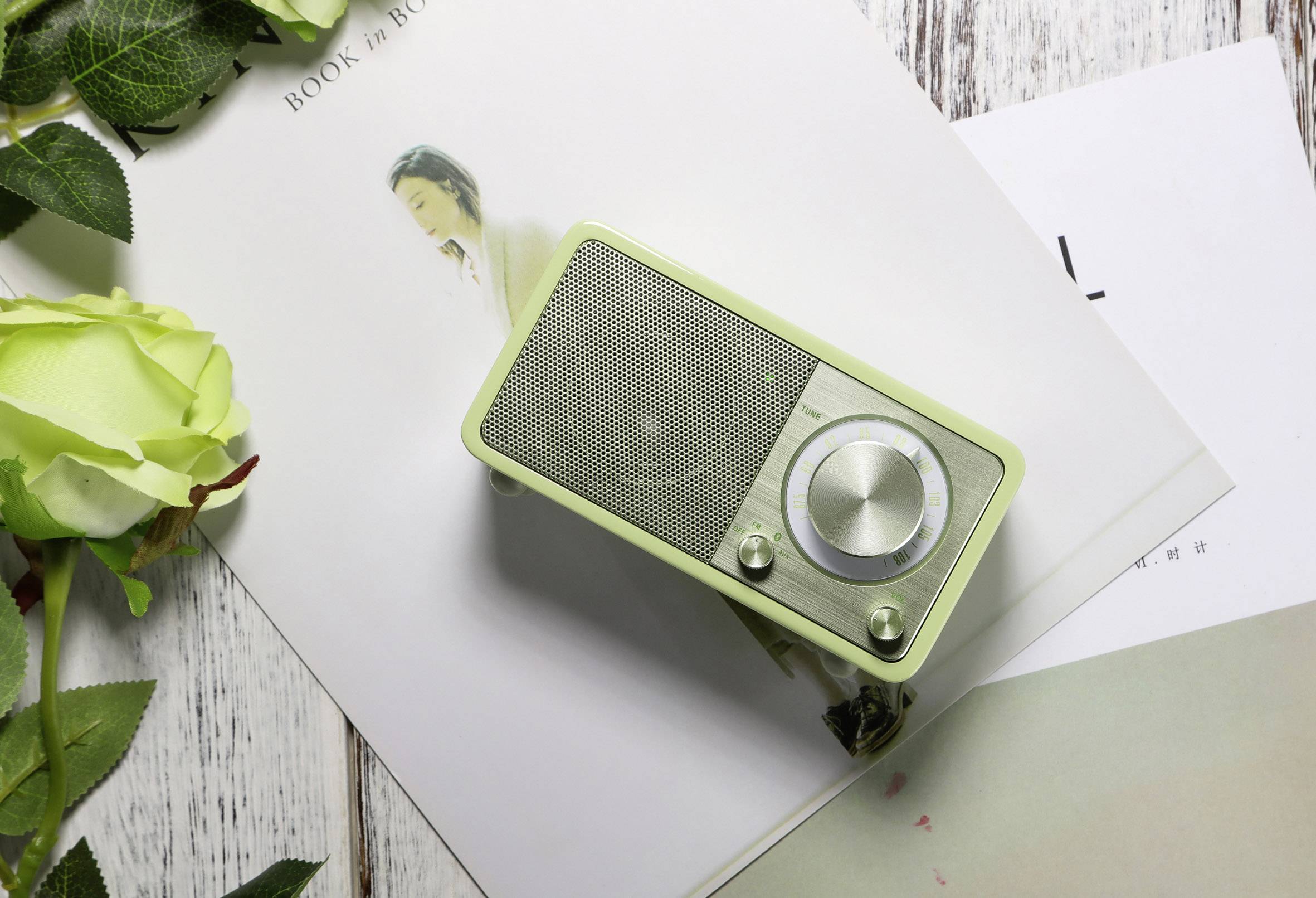 A small, green speaker sits on a wooden table. Next to the speaker are a magazine and a green rose.