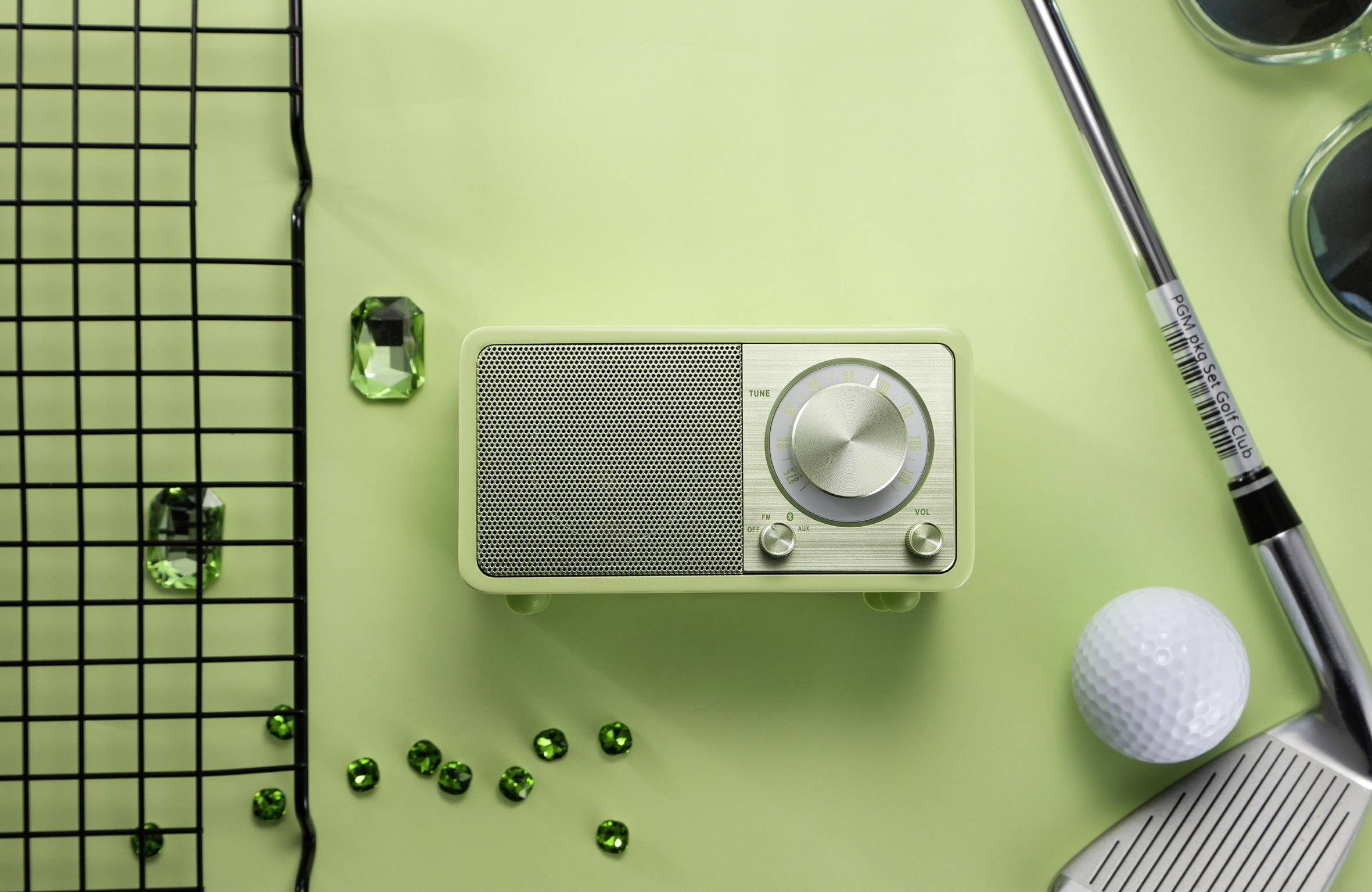 A green radio on a table with a golf ball, golf club, green gemstones, and sunglasses. Sports and leisure accessories.