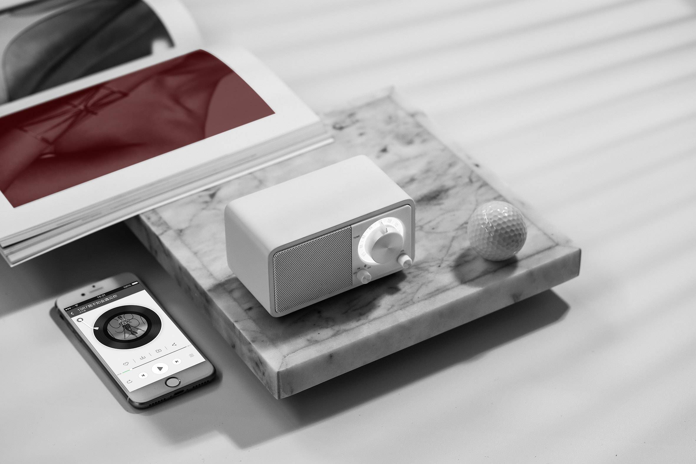 A stylish, minimalist arrangement of an open book, a speaker, a golf ball, and a smartphone on a marble tray.