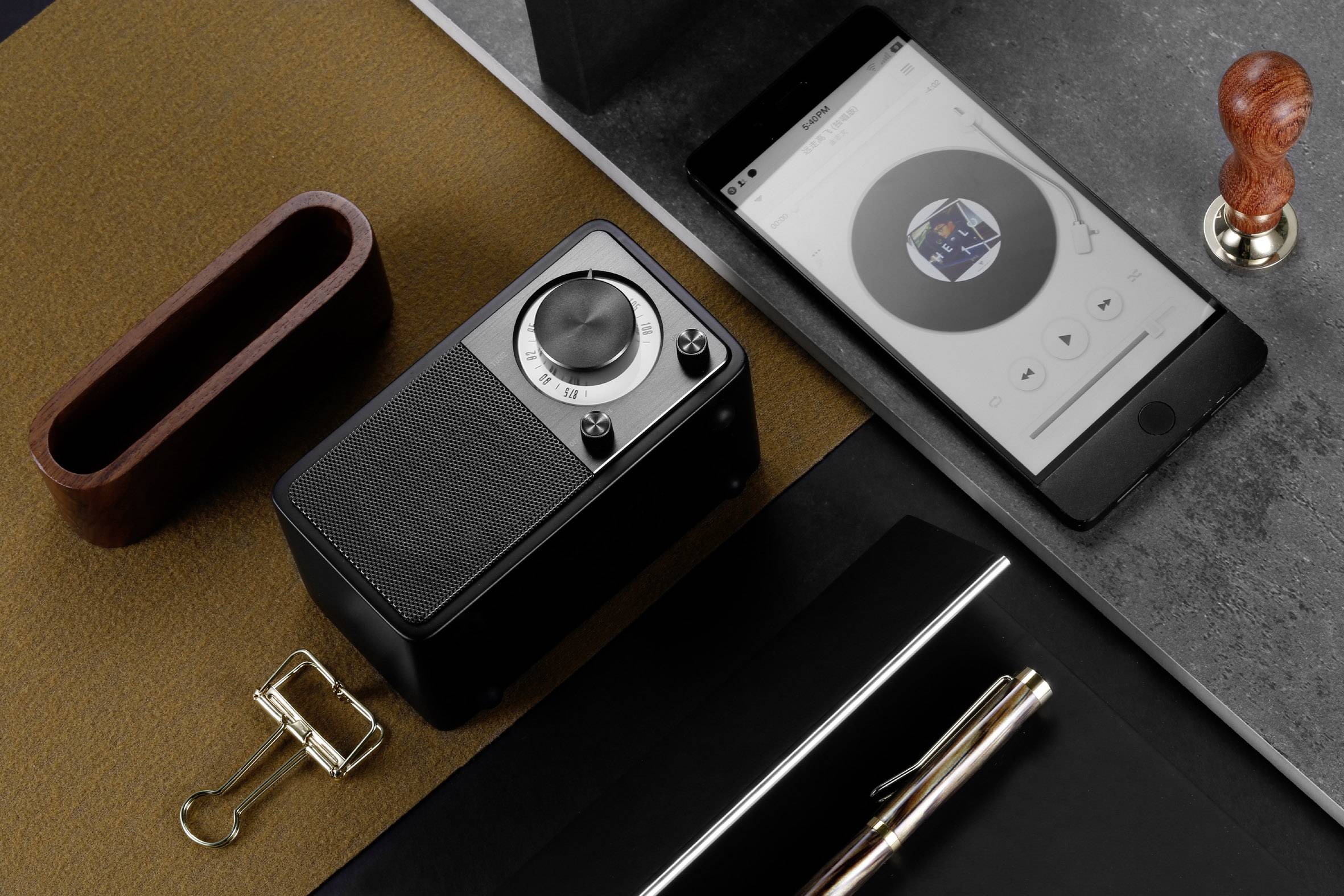 On a table, there is a speaker, a smartphone with a music app, a rubber stamp, paperclips, and a pen on a notepad.