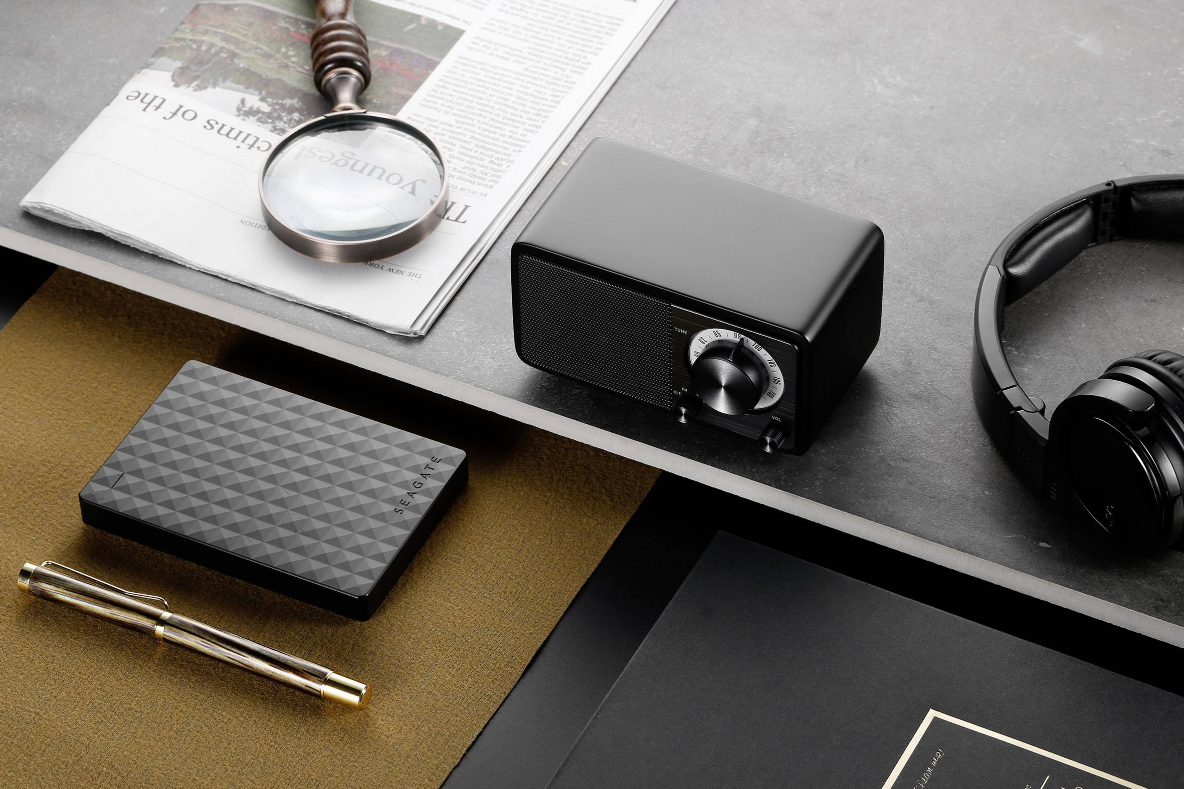 Desk with a magnifying glass on a newspaper, a black radio, headphones, a pen and a hard drive, with a black notebook beside them.