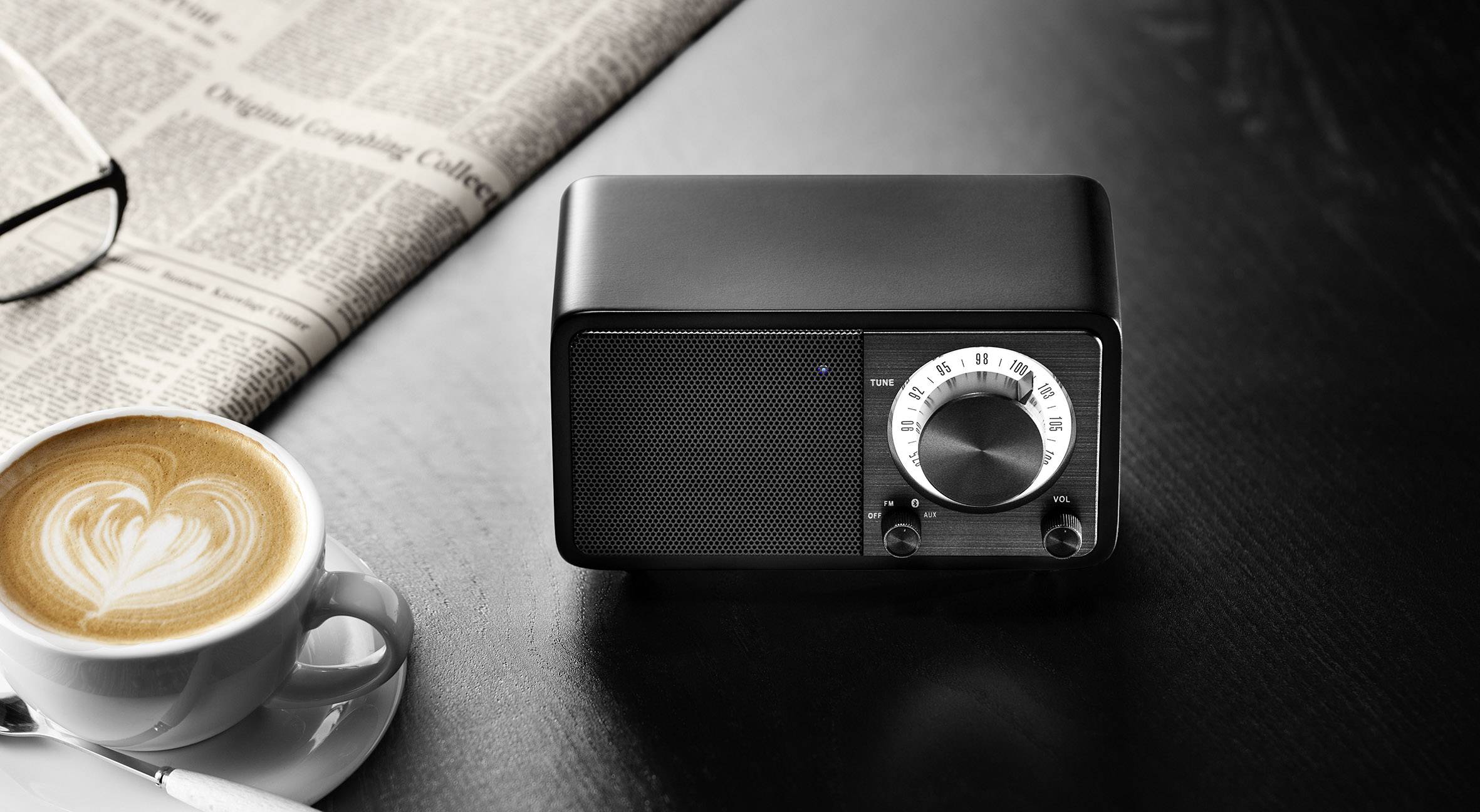 A black radio with rotary controls sits on a table beside a cup of cappuccino and a newspaper.