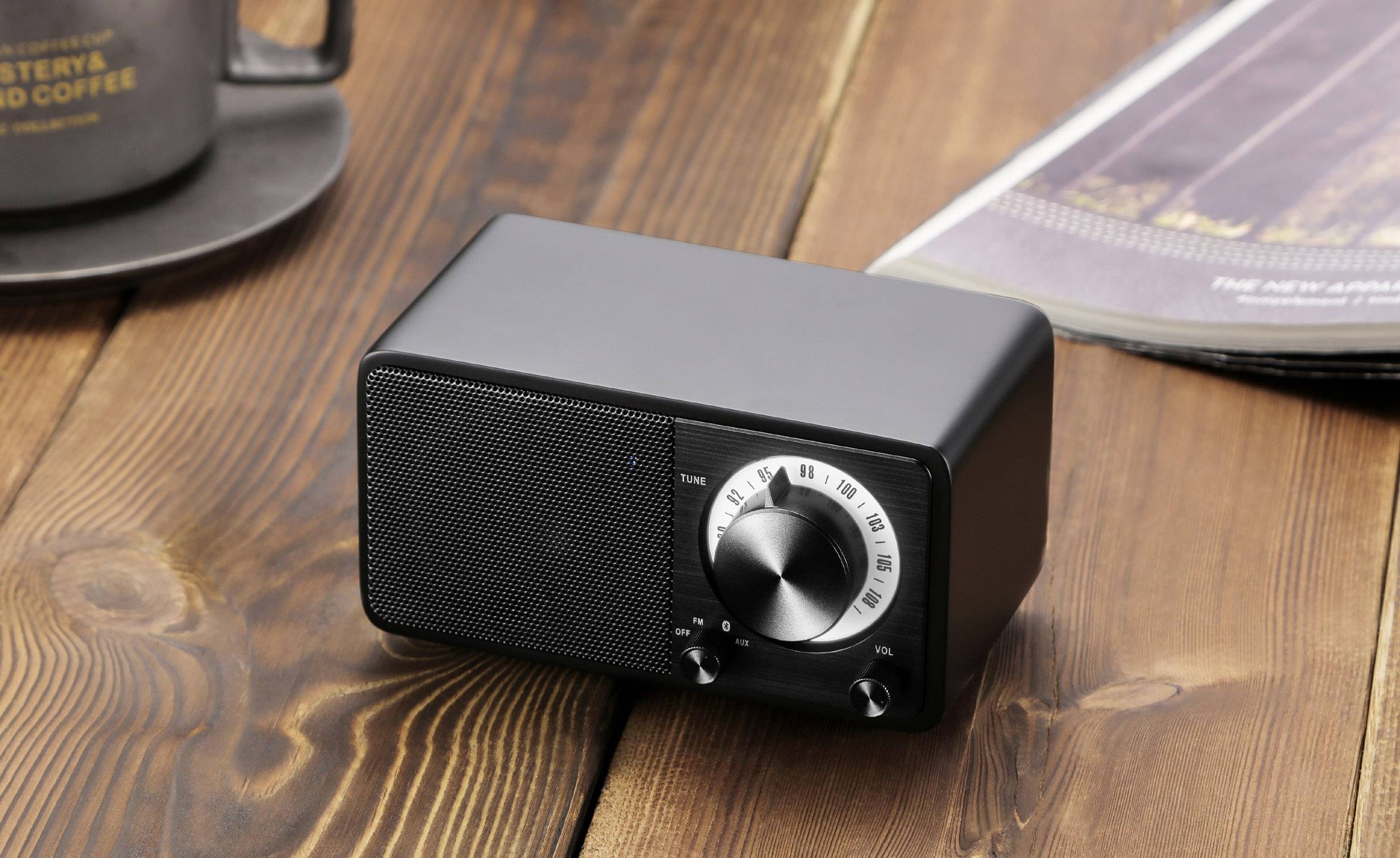 A small black speaker with rotary knobs sits on a wooden table. Next to it is a coffee cup and an open magazine.
