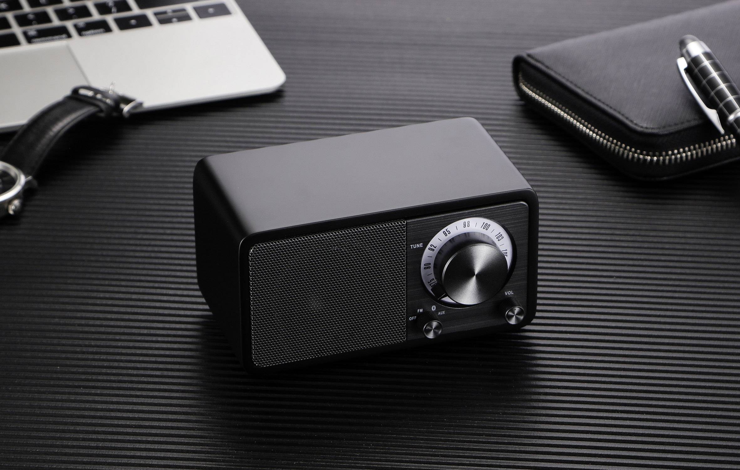 A black, rectangular speaker with a rotary knob and scale on a table. Next to it, a laptop, a black bag and a pen.