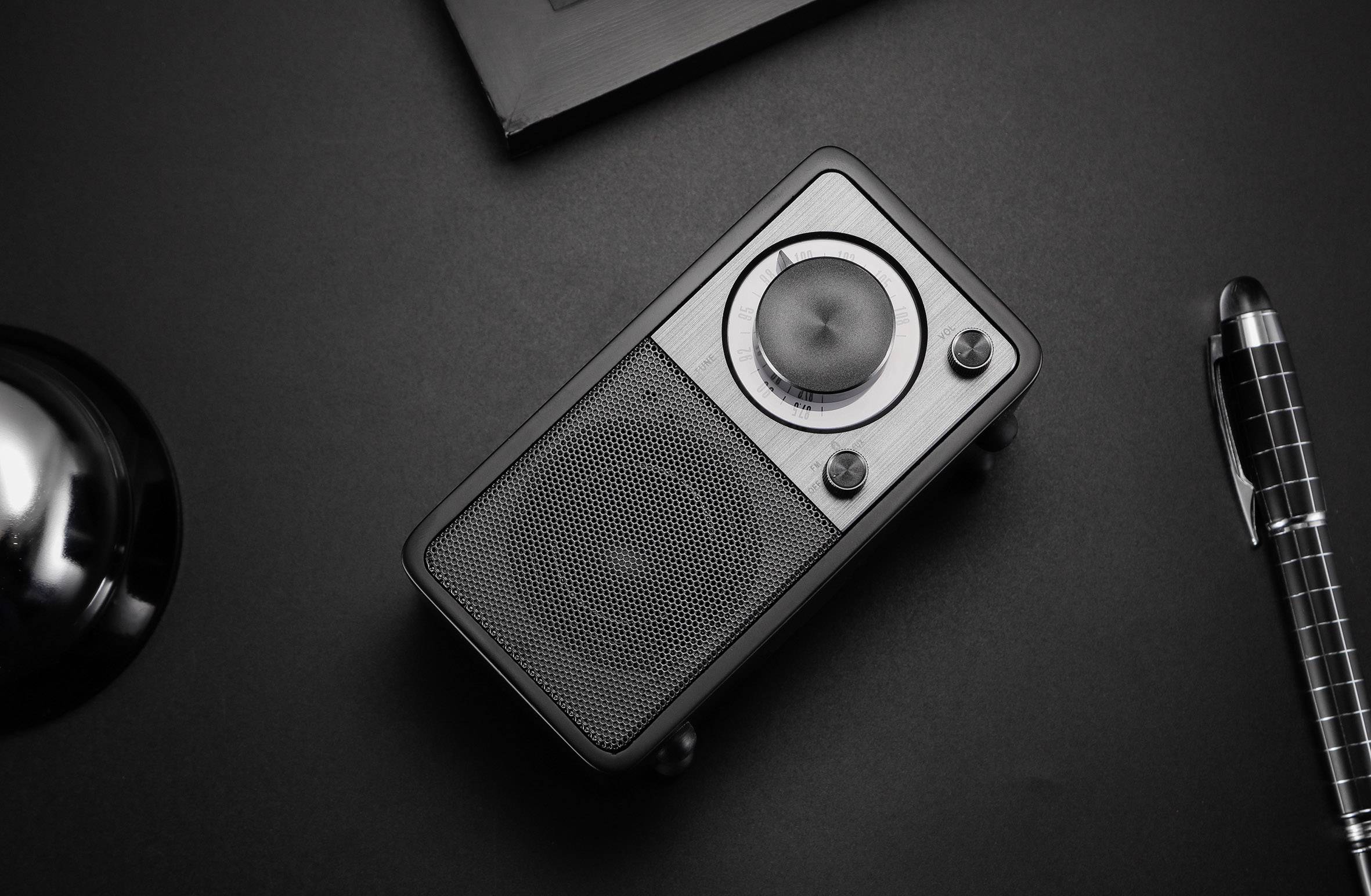 A small black portable speaker on a table. Beside it lies a black and silver pen.