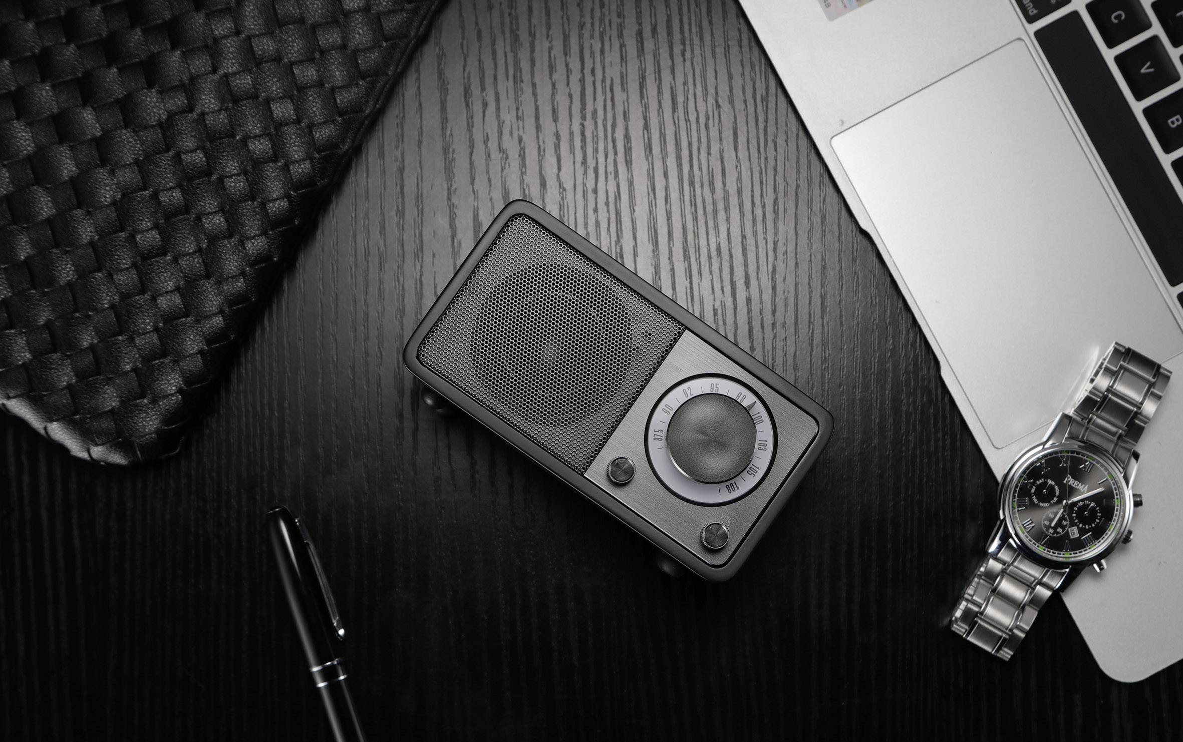 A black tabletop radio with speakers and controls sits on a dark wooden table next to a laptop, a clock, and a pen.