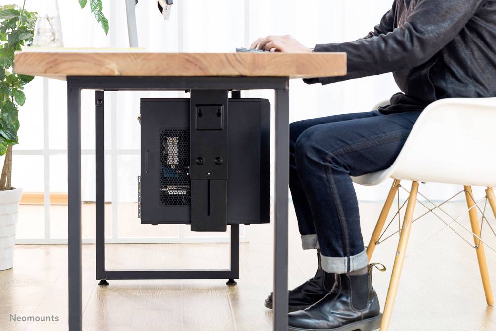 A person is working at a desk on a computer. The PC is mounted underneath the desk. Plants are visible in the background.