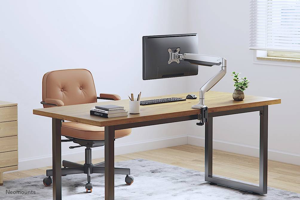 A modern desk with a brown chair, monitor on an adjustable arm, keyboard, plant, and notebook on top, in a bright room.