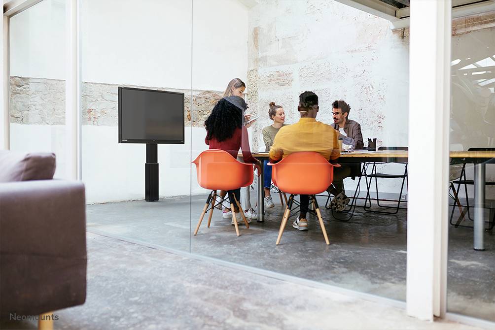 A group of five people are sitting around a table in a modern conference room. A large screen is hanging on the wall.