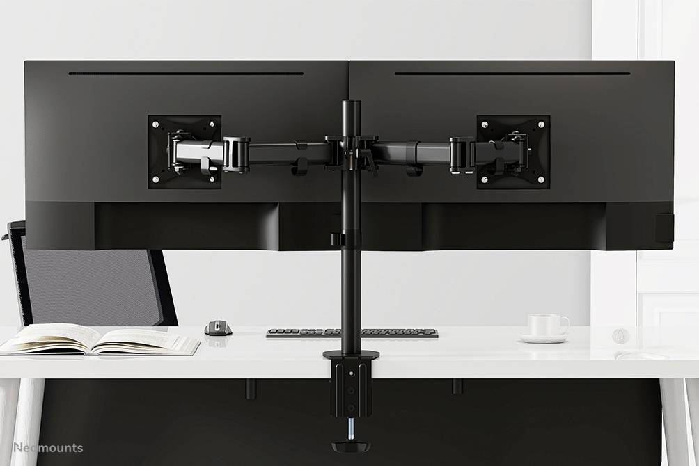Two black monitors on a mount, positioned on a desk. Beside them lie an open book, a mouse and a cup.