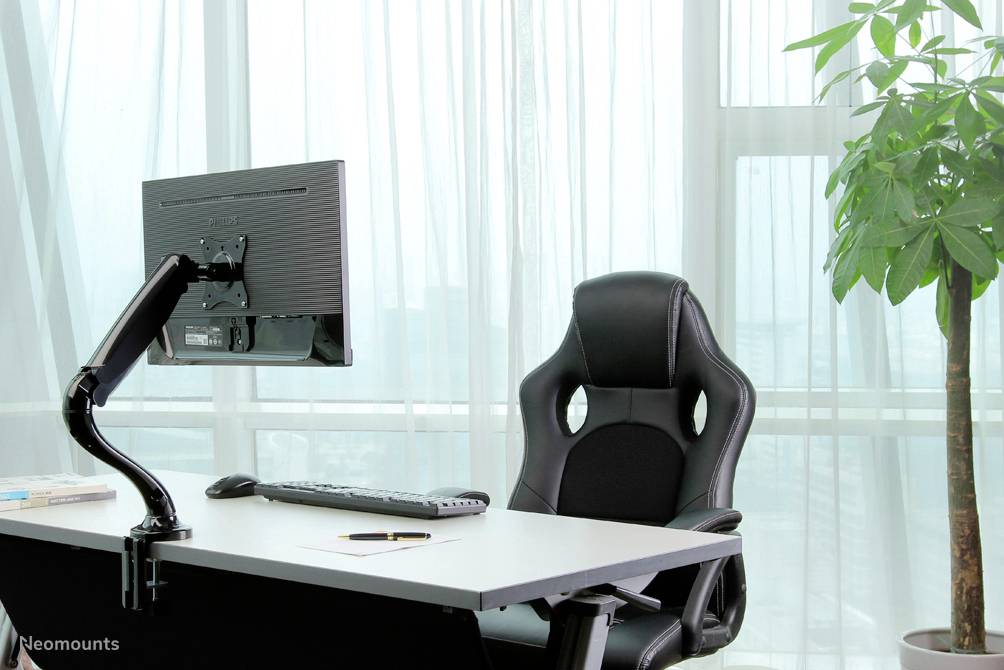 A modern office space with a desk, screen, keyboard and ergonomic chair. A large plant stands to the right by the window.