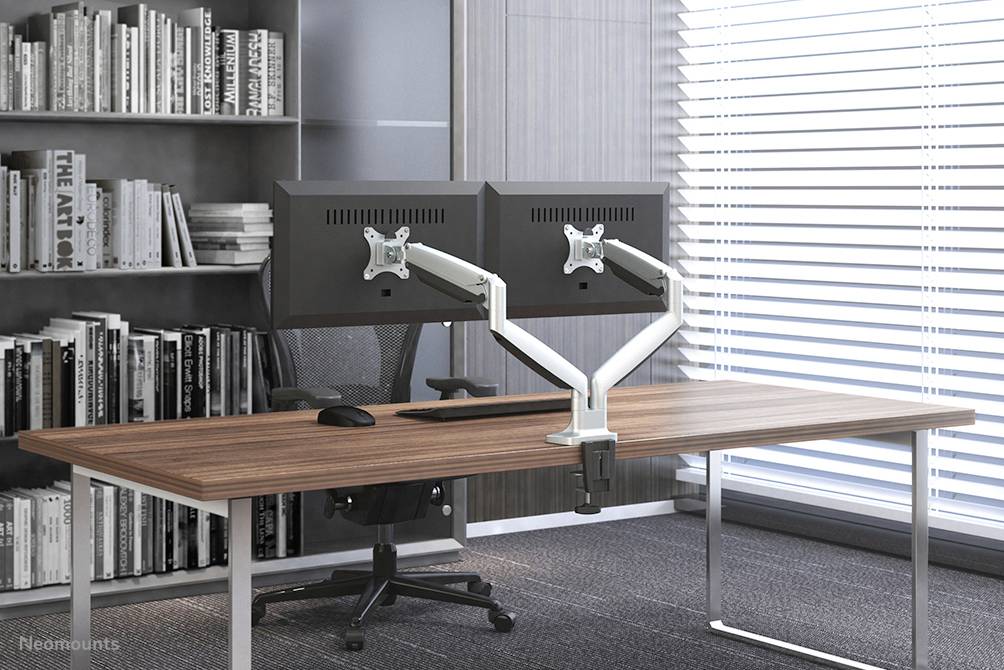 A modern desk with two monitors on mounting arms, a black office chair, and a bookshelf in the background.