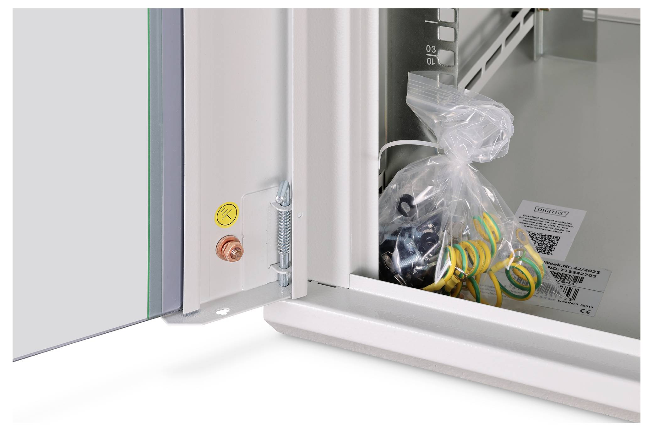 An open cabinet door showing a plastic bag containing colored electrical components and a manual inside. A grounding bolt is visible on the door.