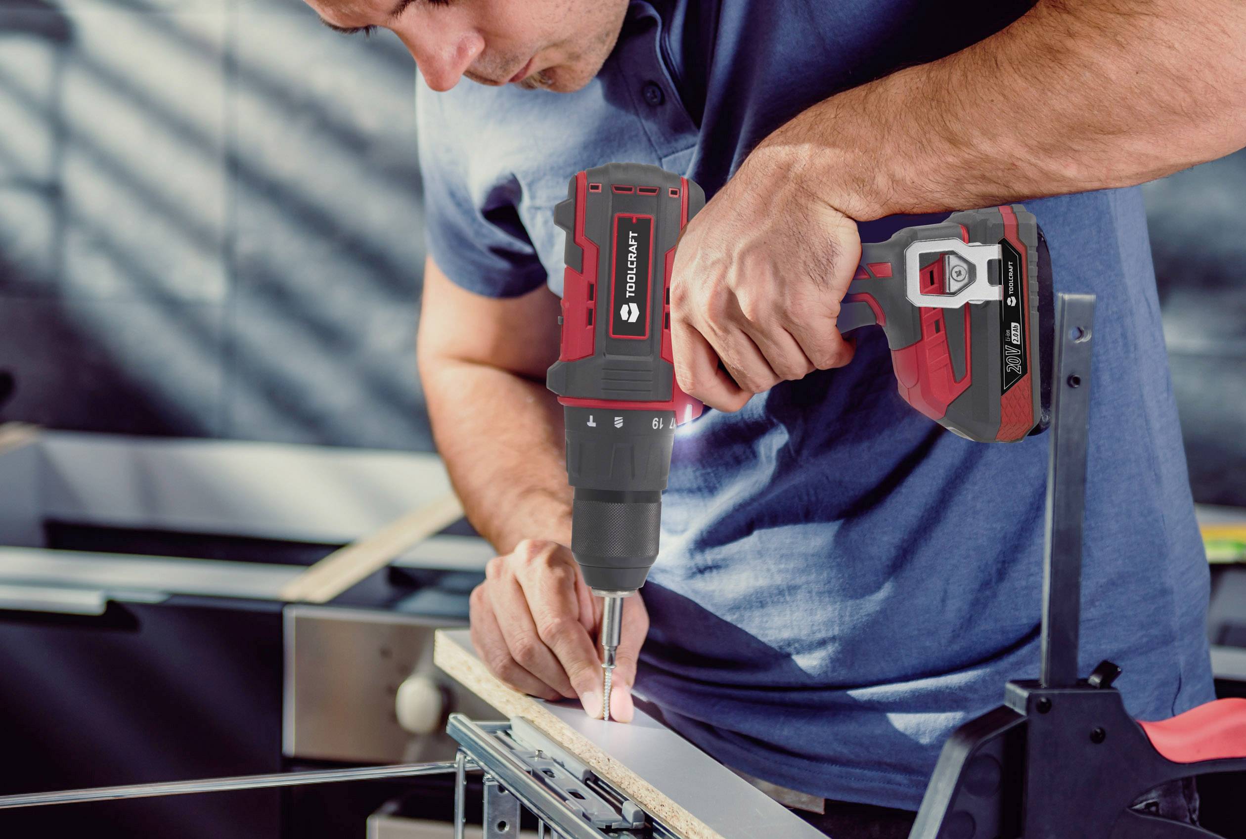 A man is using a cordless drill to turn a screw into a piece of wood in a workshop environment.