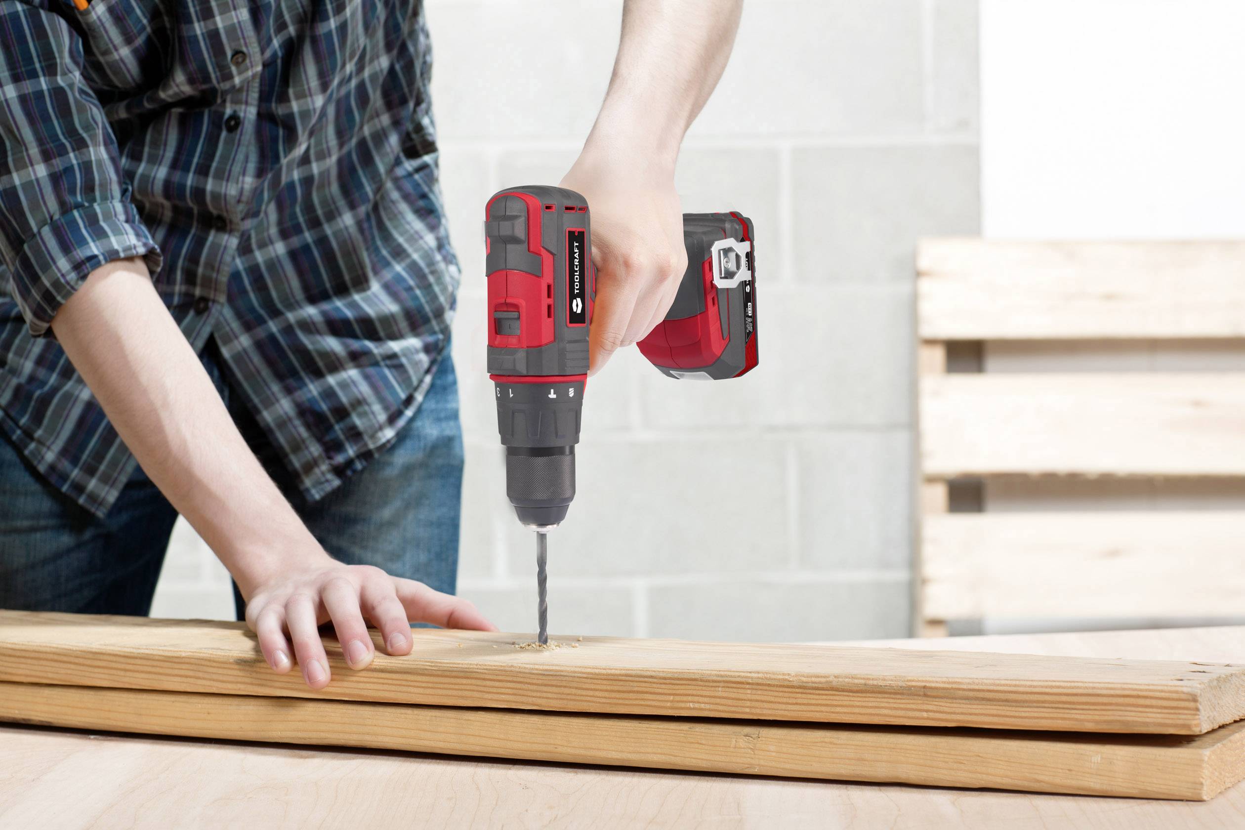 A person wearing a checked shirt is drilling a hole in a wooden board with a red cordless drill in a workshop setting.
