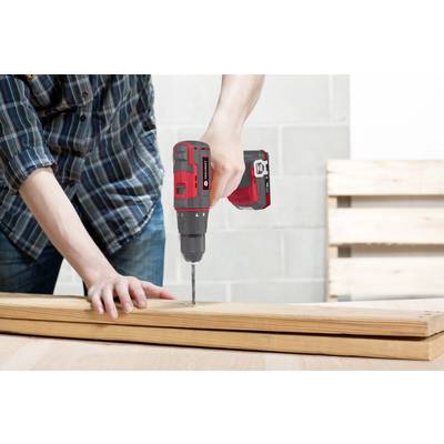 A person wearing a checked shirt is drilling a hole in a wooden board with a red cordless drill in a workshop setting.