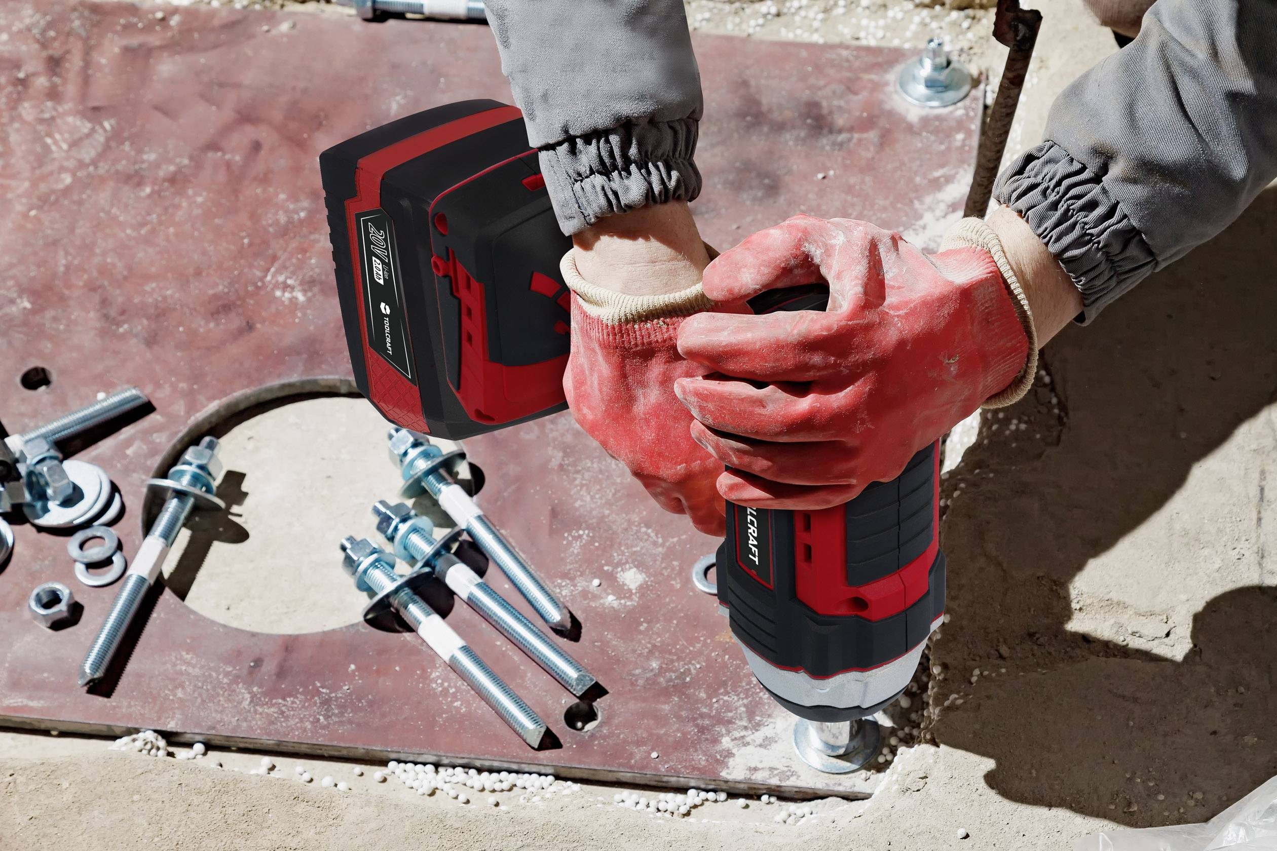 A person wearing red gloves is holding a cordless drill and drilling holes into a metal plate, with screws and nuts lying nearby.