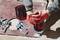 A person wearing red gloves is holding a cordless drill and drilling holes into a metal plate, with screws and nuts lying nearby.