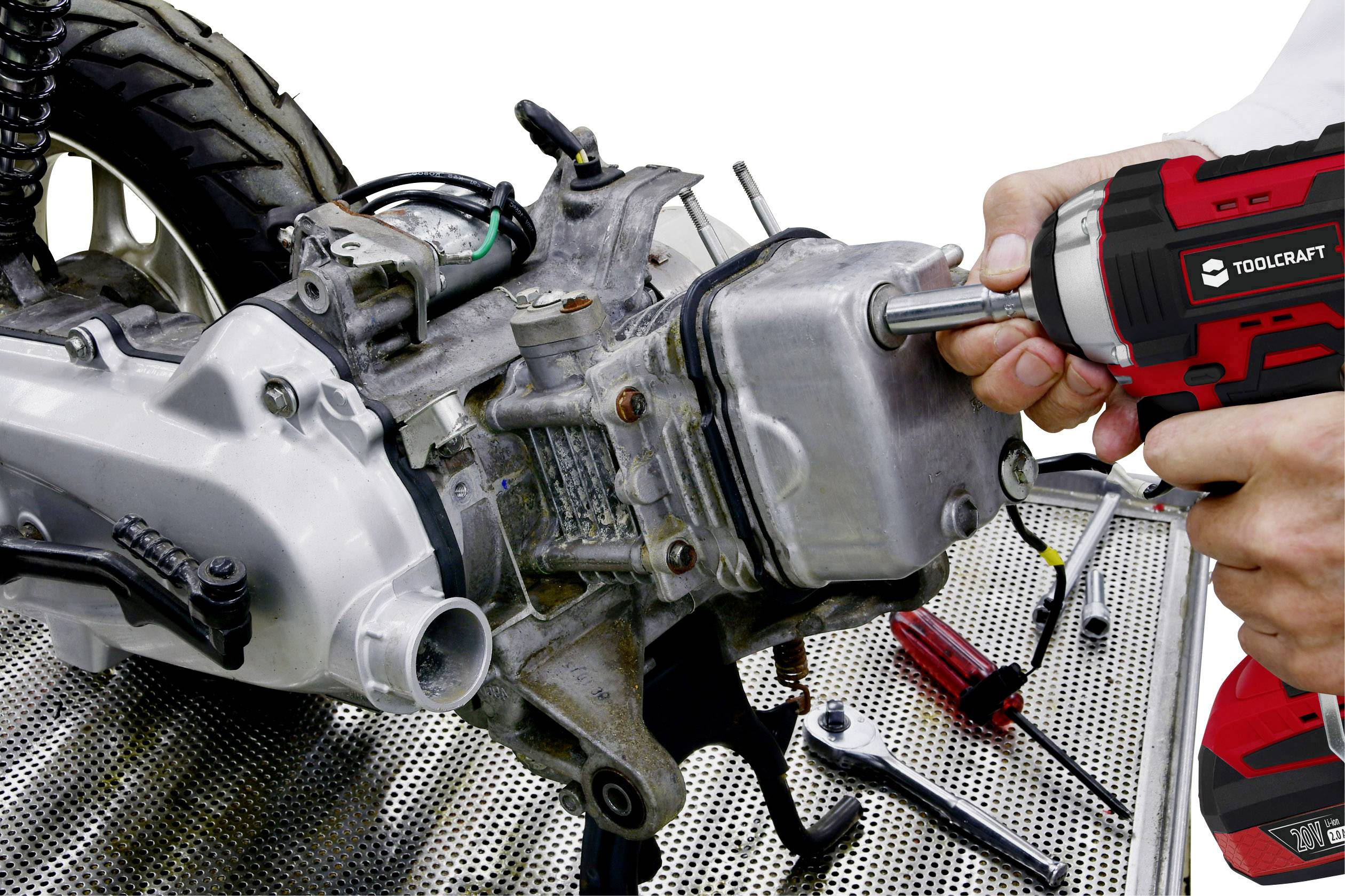 Two hands are working with an electric screwdriver on a motorcycle engine on a workshop bench. Various tools are visible.