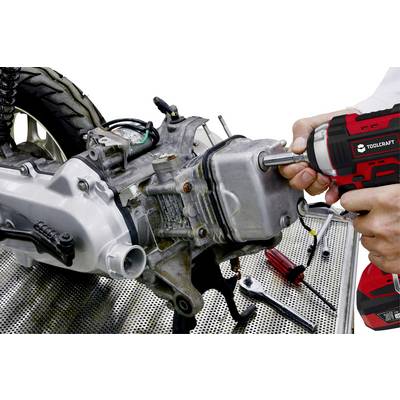 Two hands are working with an electric screwdriver on a motorcycle engine on a workshop bench. Various tools are visible.
